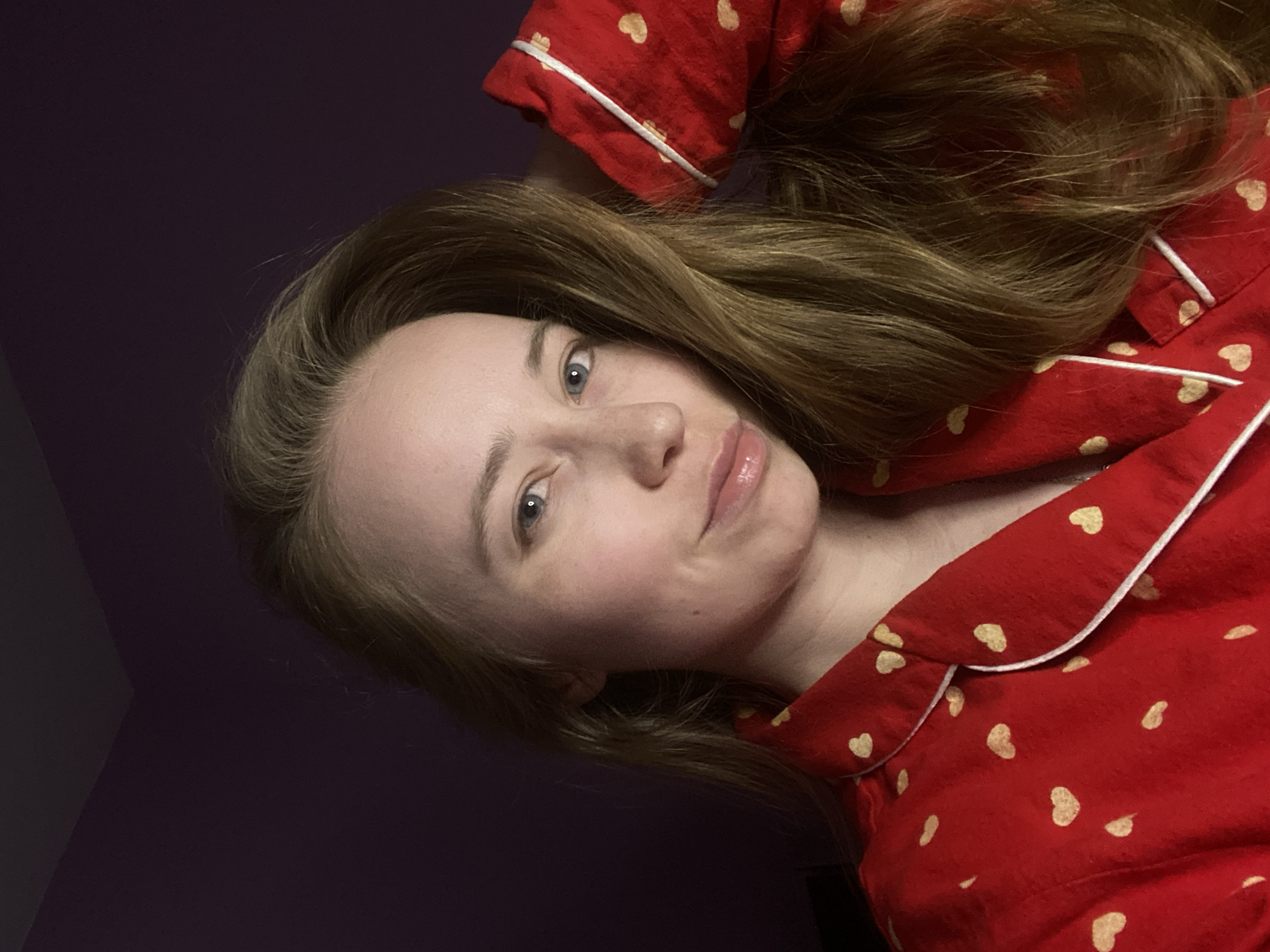 A woman with light skin and long, light brown hair is seen from the chest up, lying down and looking towards the camera. She has blue eyes and is wearing red pajamas with a white trim and yellow hearts on them. The background is dark, possibly a wall or room in low light. There are no indications of the image being taken in Saint Petersburg, Russia.