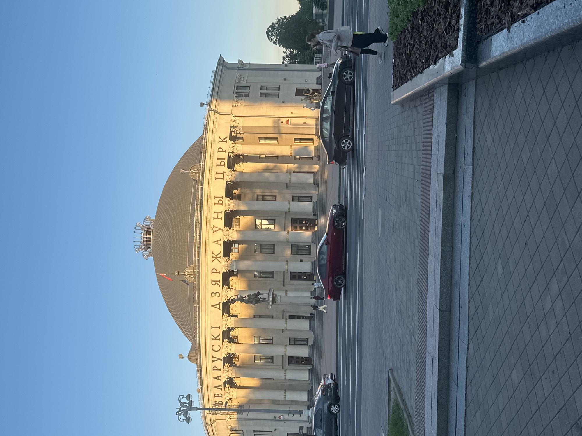The image depicts a street scene dominated by a grand, classical-style building with a large domed roof, bathed in the warm light of a low sun. The sky above is clear and blue.
The prominent building is identified by Cyrillic text inscribed along its facade, which reads "БЕЛАРУСКІ ДЗЯРЖАЎНЫ ЦЫРК" (Belarusian State Circus). Its front features numerous tall, ornate columns and sculptural elements, with windows visible between them. A textured dome tops the structure, and a red flag is visible on a flagpole near the roofline.
In front of the building, a paved road with white lane markings runs parallel to the facade. Several cars are present; a black sedan is parked close to the building, and a red sedan is further down the street, with another dark vehicle partially visible. A wide, tiled sidewalk separates the road from a raised planter filled with dark mulch and some greenery.
A few people are visible in the scene. One person is walking on the sidewalk near the black car, and other figures can be seen near the building's entrance in the distance.
The long shadows cast by the columns and vehicles suggest that the photograph was taken during either early morning or late afternoon. The overall setting is an urban environment on a clear, sunny day in Minsk, Belarus, capturing a typical moment in front of a significant cultural landmark.