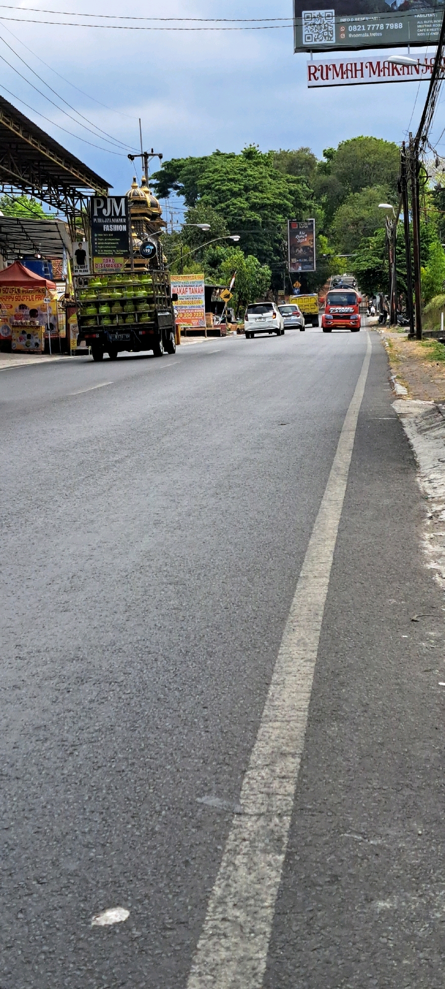 The image depicts a street scene in Pandaan, Indonesia, on a cloudy day. A truck carrying numerous green gas cylinders is driving down the road. In the foreground, the asphalt road is visible with a white line marking the lane. To the left of the road, there are several street vendors with colorful stalls and signage. One sign reads "PJM FASHION". Further down the road, other vehicles, including cars and a red truck, are also present. Trees and foliage line the right side of the road, and a few streetlights are visible. A large billboard advertising "RUMAH MAKINJAYA" is seen in the upper part of the image. The overall impression is a bustling street with commercial activity.
