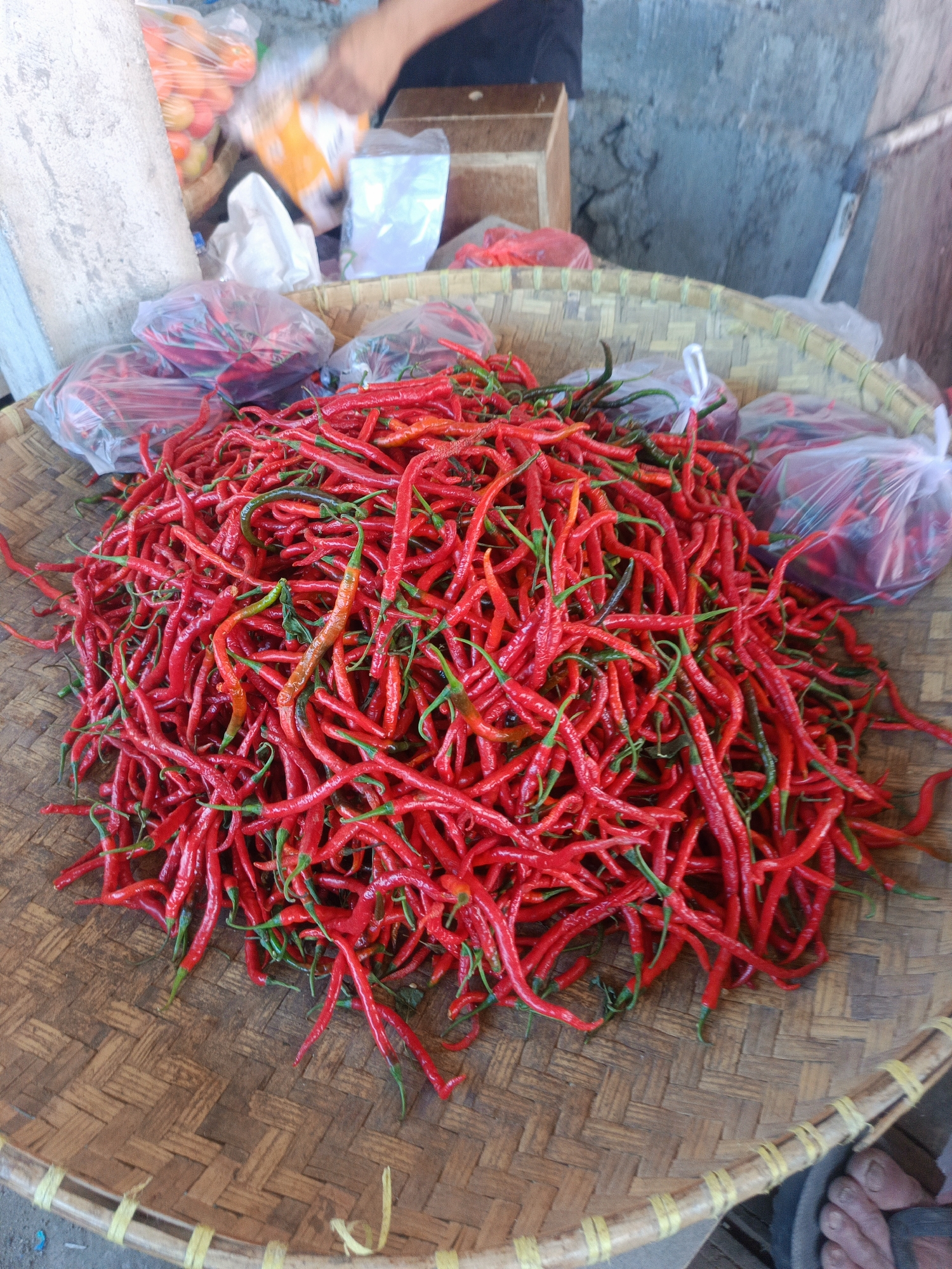 This is a close-up, overhead, eye-level shot of a large pile of red chilies in a woven bamboo basket. The chilies are long and slender, with some showing green stems. They are piled high in the basket, with some chilies spilling over the edge. The basket is placed on a textured surface, possibly a wooden stall. In the background, there are out-of-focus plastic bags containing produce, and a concrete wall. A person's hand is visible in the upper left corner, reaching towards the produce. The scene appears to be at a market, likely in Gorontalo, Indonesia, given the context.