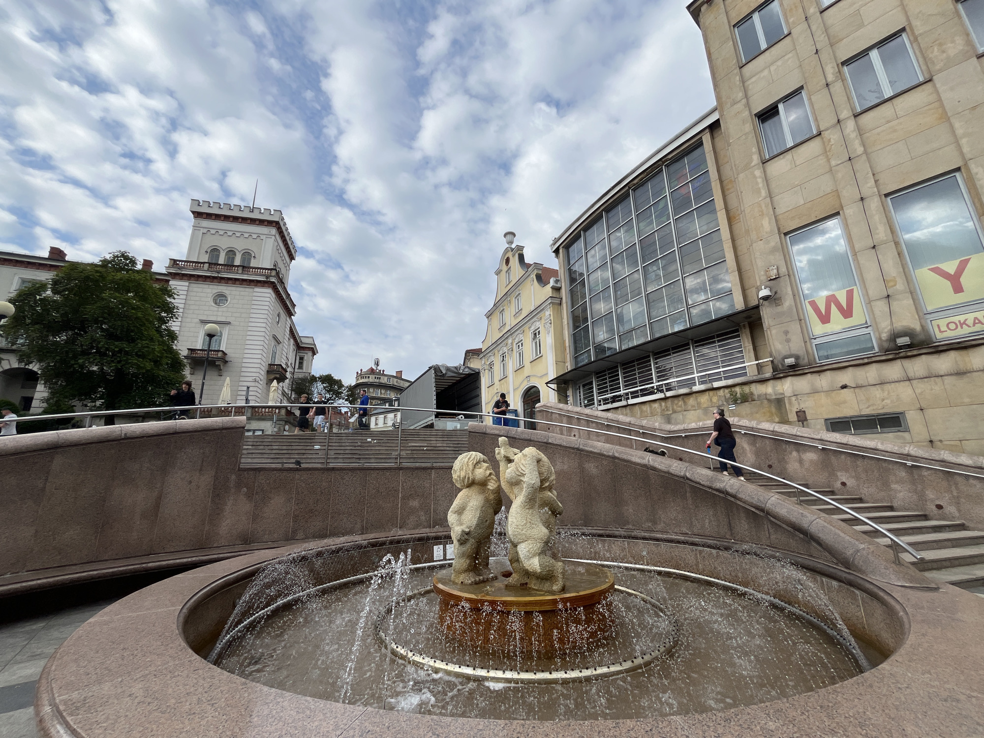 In the foreground, a circular fountain with rippling water features a small sculpture of two children. Behind the fountain, a tiered stone staircase ascends, with a person walking up the stairs on the right side of the image. The stairs are bordered by ornate stone walls and metal railings. In the background, several buildings are visible. To the left, a light-colored building with a tower and battlements stands next to a lush green tree. In the center, a yellow building with multiple windows is situated. To the right, a modern building with a glass facade and large windows, some displaying the letters "W" and "Y" and the word "LOKAL," dominates the scene. The sky above is filled with scattered clouds. The overall impression is an urban plaza with architectural elements and public art.