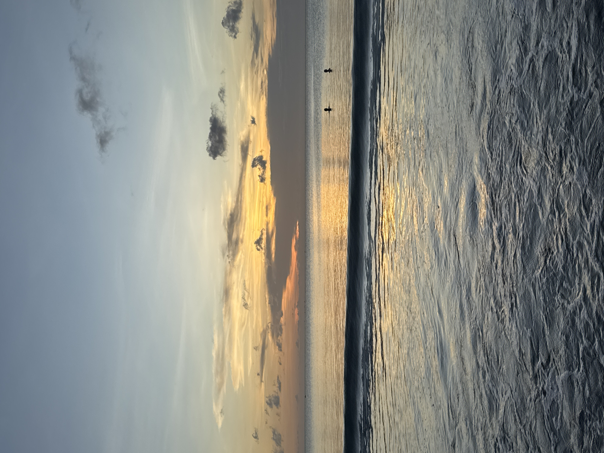 The image depicts a scene at Jimbaran Beach, Indonesia, at sunrise or sunset. The sky on the left is a gradient of blues and yellows, with scattered clouds. The ocean is calm and reflects the light, creating a golden sheen. There are two figures in the water. A wave is visible along the shoreline. The sand appears textured and wet.