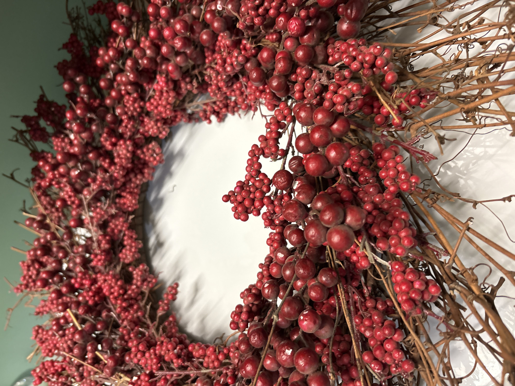 This media file is a close-up, vertically oriented, outdoor photograph of a wreath made of red berries and brown twigs. The wreath is circular and is arranged on a surface that appears to be a white wall or backdrop, with a subtle shadow cast by the wreath. The lighting suggests an indoor setting, possibly a home or a craft store. The wreath is densely packed with clusters of small, red berries and interspersed with thin, dried brown twigs and vines. Some of the berries are glossy, while others have a matte finish, indicating they might be artificial. The texture of the berries is varied, with some appearing smooth and round, and others slightly elongated or clustered together. The overall impression is one of a rustic, decorative item, likely intended for holiday or seasonal display. There is no visible text in the image. The focus is entirely on the detailed texture and arrangement of the wreath's components. The image does not depict any people, animals, or specific environmental conditions like weather. The implied location of Bradenton, United States, provides a general geographical context but does not offer specific visual cues within the image itself.