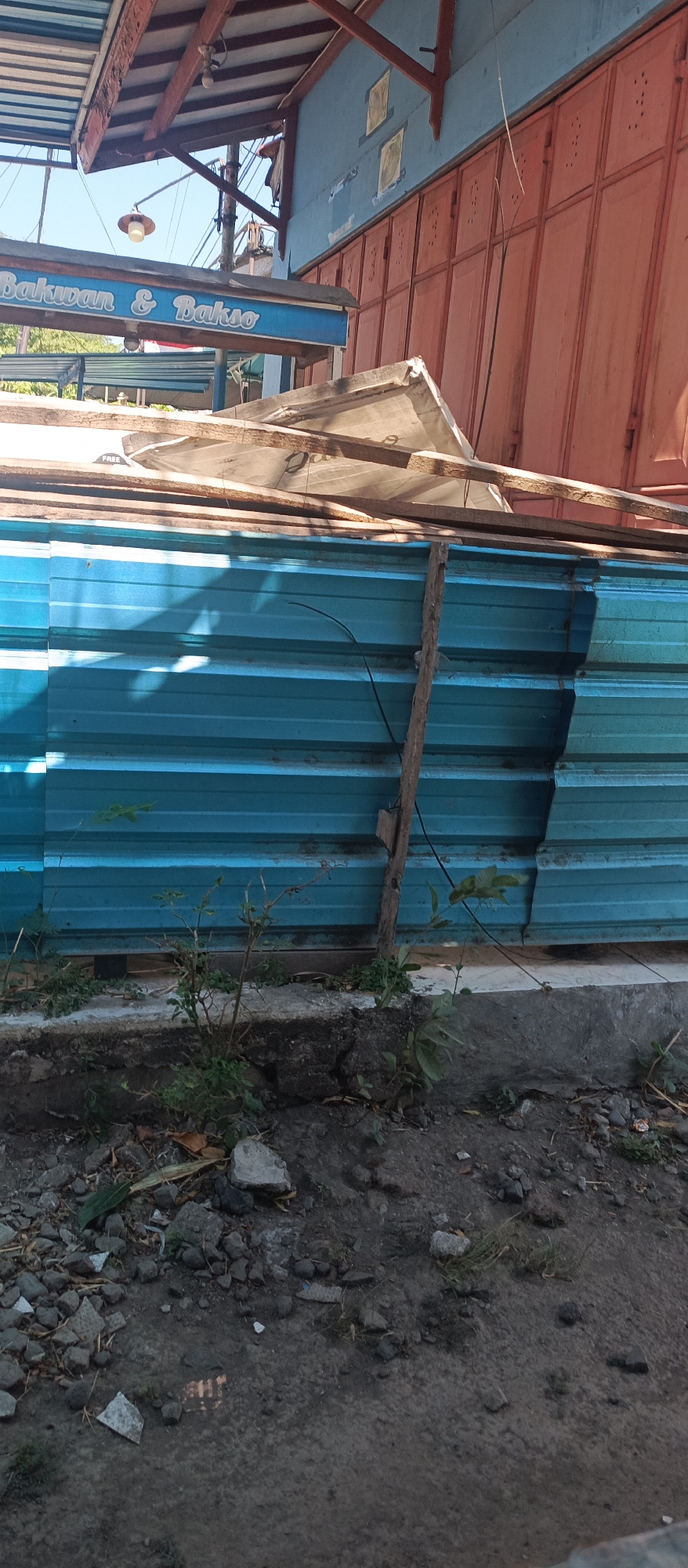 A blue corrugated metal barrier is stacked in front of a building with orange folding doors. A wooden beam supports the barrier. A light fixture hangs from the awning of the building. The building has a sign that reads "Bakwan & Bakso". The ground in front of the barrier is covered with debris and small plants. The scene appears to be in Jatisari, Indonesia.