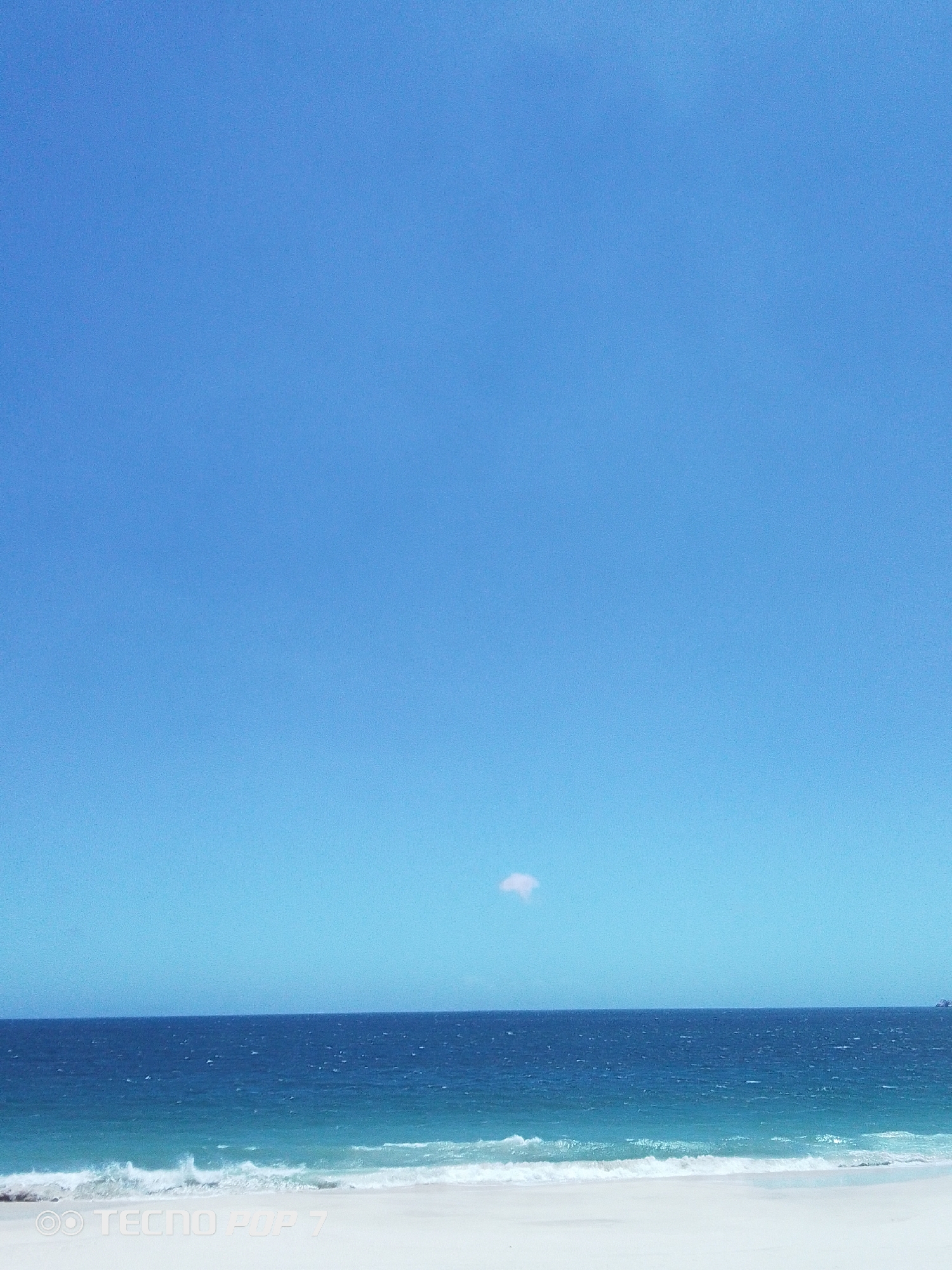 The image presents a serene coastal landscape captured in Pedro González, Venezuela. The scene is dominated by a vast, clear blue sky, occupying the upper two-thirds of the frame. A single, small, faint white cloud is visible in the sky near the horizon line.
The horizon is sharply defined, separating the expansive sky from the ocean. The ocean itself displays varying shades of blue, with deeper blues in the distance transitioning to lighter, turquoise hues closer to the shore. Gentle, white-capped waves are seen breaking onto a pristine white sandy beach, which fills the foreground. In the far right distance, a small, dark, indistinct shape, possibly a distant island or landmass, is barely visible.
No people or significant man-made objects are present, emphasizing the natural beauty and tranquility of the setting. The bright, clear blue sky and the quality of light indicate a sunny day. The overall impression is one of calm and natural grandeur.
In the bottom left corner of the image, the watermark "OO TECNO POP 7" is visible.