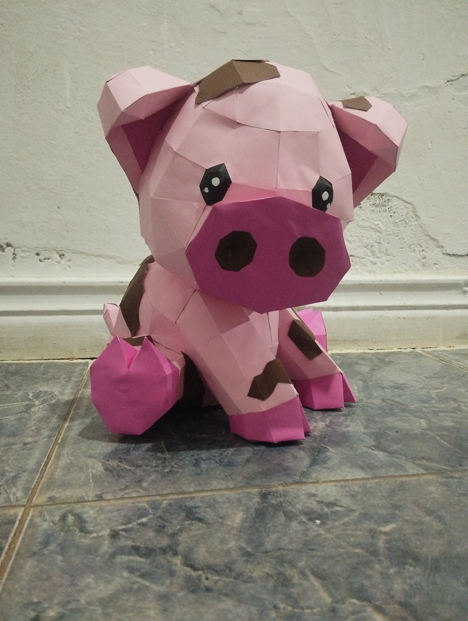 A low-angle, full shot depicts a papercrafted pig figurine sitting on a tiled floor against a white wall. The pig is primarily pink with brown spots. Its head features prominent, large, angular ears, black eyes, and a dark pink snout with dark brown nostrils. The pig's body is constructed from various polygonal shapes. The figurine sits squarely on the floor tiles, which are predominantly gray-blue and show a veined pattern. The background wall is a stark white with a slightly textured surface. The location is El Valle del Espíritu Santo, Venezuela. No people are present.