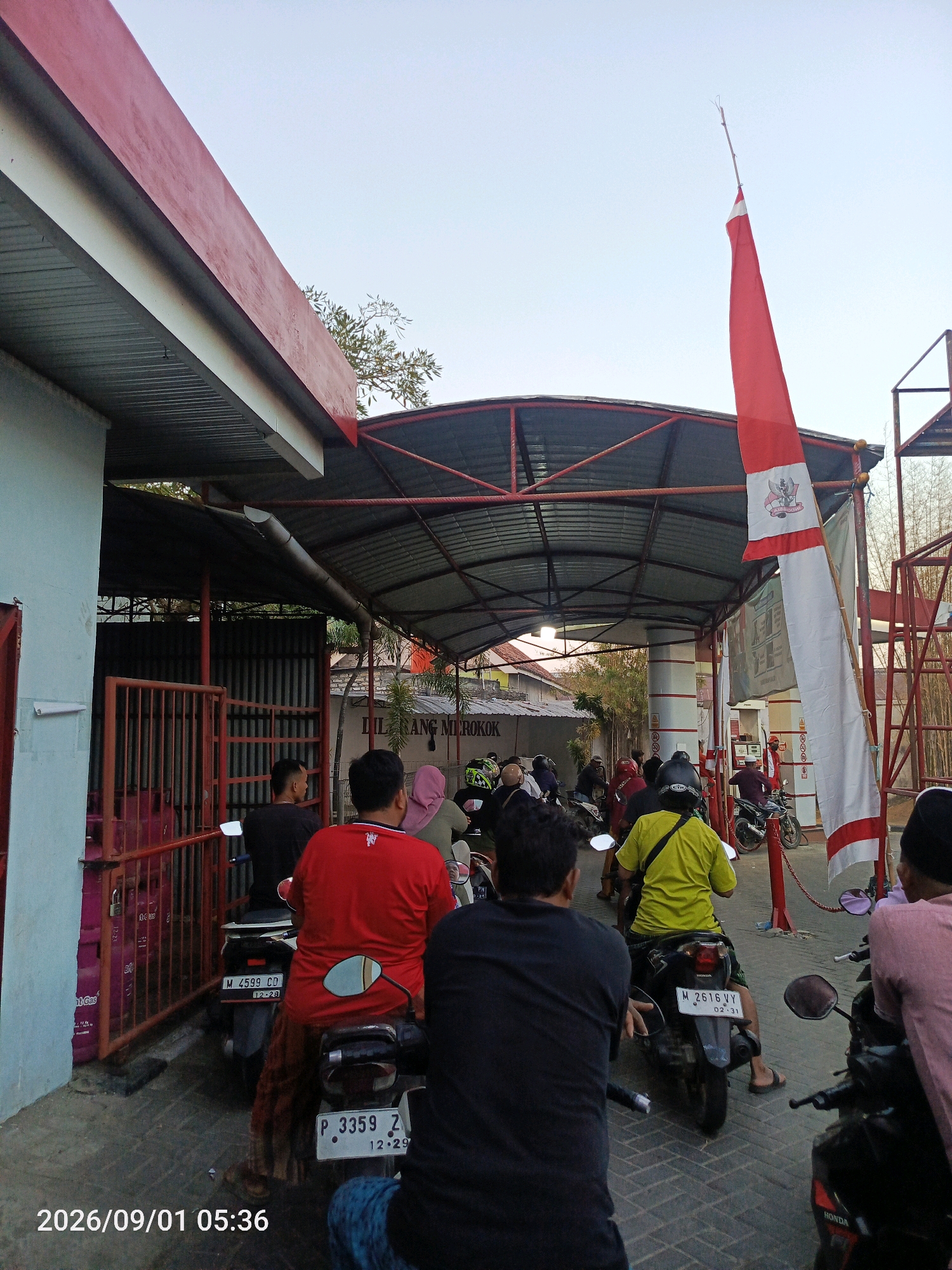 The image depicts an outdoor scene, likely at a fuel station, with several people on motorcycles. The setting appears to be in Gladak Anyar, Indonesia, indicated by the timestamp and the presence of Indonesian cultural elements like the red and white flag. In the foreground, several individuals are seated on their motorcycles, facing away from the camera. One person in a red shirt is prominent, with a motorcycle license plate visible. Further back, a line of motorcycles and riders extends into a covered area. The architecture suggests a functional structure, possibly a gas station canopy, with corrugated metal roofing supported by columns. The environment includes a clear sky, some vegetation, and a prominent red and white flag flying from a pole. A sign that reads "DILARANG MEROKOK" (No Smoking) is visible on a building in the background. The overall atmosphere is calm and everyday, with people engaged in routine activities.