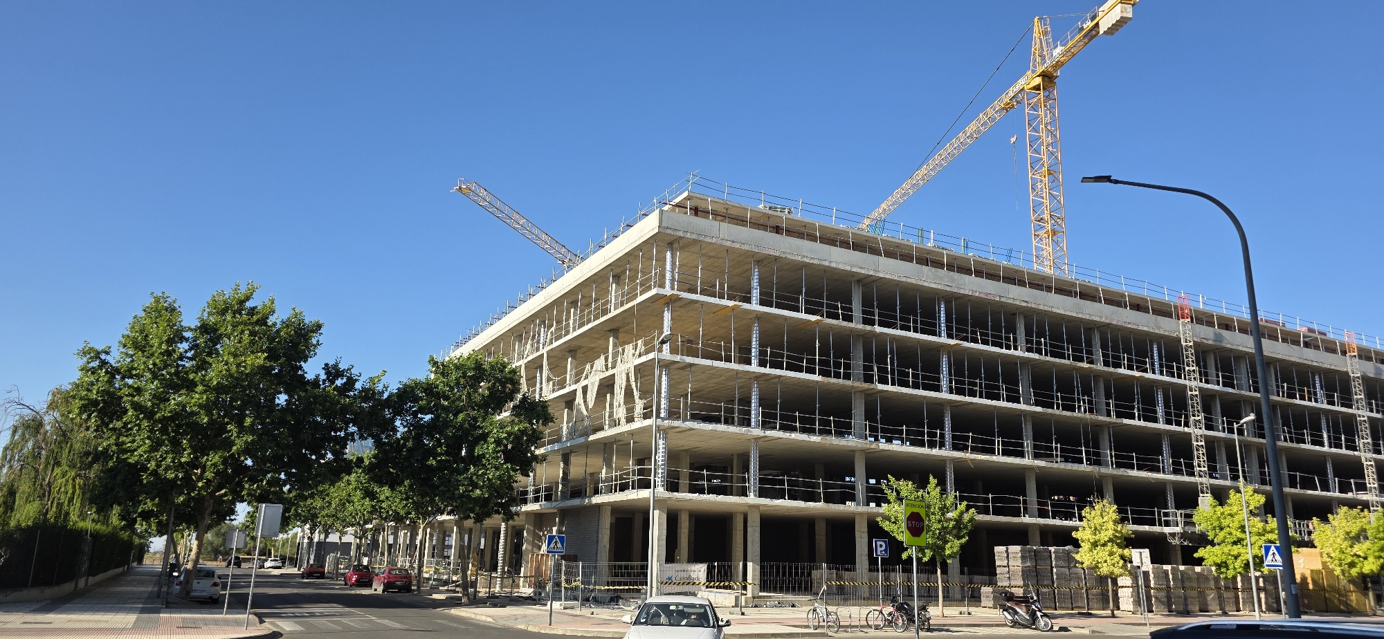 An outdoor, eye-level, wide shot captures a large, multi-story building under construction in Huesca, Spain. Two yellow construction cranes are visible against a clear blue sky. The building, composed of concrete and scaffolding, has multiple open floors with visible support columns and railings.
In the foreground, a paved street curves to the left, with a pedestrian sidewalk to the right of the street. Several green trees line the sidewalk, with one particularly large tree dominating the left side of the frame. Red and silver cars are parked or driving on the street. A white car is visible on the sidewalk near the lower right corner of the image.
Further down the sidewalk, a street lamp with a curved arm stands tall. Signs on poles, including a blue pedestrian crossing sign and a green sign with "ATENCION STOP," are present. Stacked materials, likely construction supplies, are visible on the right side of the sidewalk. A parked scooter is also present on the sidewalk. The scene suggests a sunny day in an urban or suburban environment undergoing development.