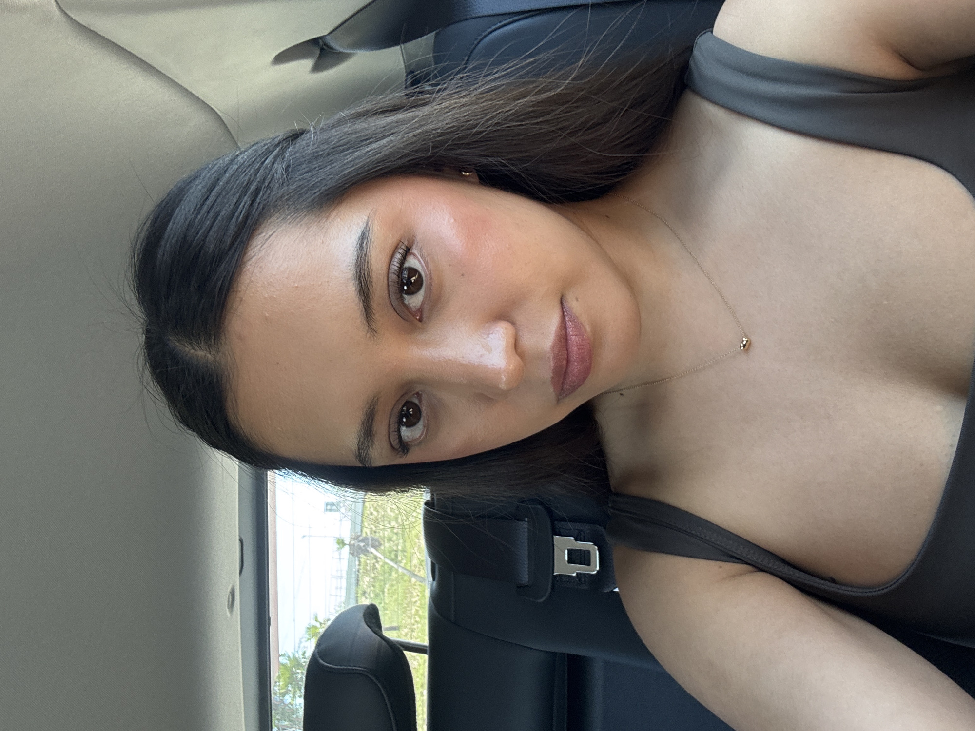 The image captures a close-up portrait of a young woman, likely in her 20s or early 30s, situated inside a car. She is looking directly at the camera with a calm, relaxed expression, possibly a soft smile. She has long, dark brown hair, dark eyes, and a natural makeup look featuring subtle blush and nude-pink lipstick. She is wearing a dark grey or charcoal-colored top with wide straps, and a delicate gold necklace with a small pendant is visible around her neck.
The setting is the interior of a vehicle, possibly the back seat, as evidenced by the dark upholstery of the seat and a silver seatbelt buckle visible near her shoulder. Bright daylight illuminates the scene, suggesting it is daytime with clear weather. Through the car window, elements of the outdoors such as green foliage and hints of distant buildings can be discerned. The overall impression is a casual, personal photo taken within a car. There is no visible text in the image. The location context for this image is San Bernardino Tlaxcalancingo, Mexico.