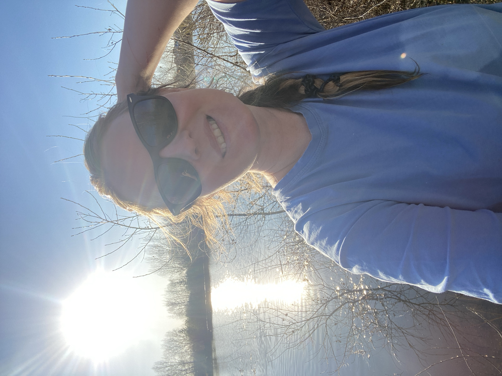 A woman wearing sunglasses and a light blue t-shirt is taking a selfie outdoors. The sun is bright, reflected on a body of water behind her. There are bare trees and brush in the background. Location information is not included in the image.