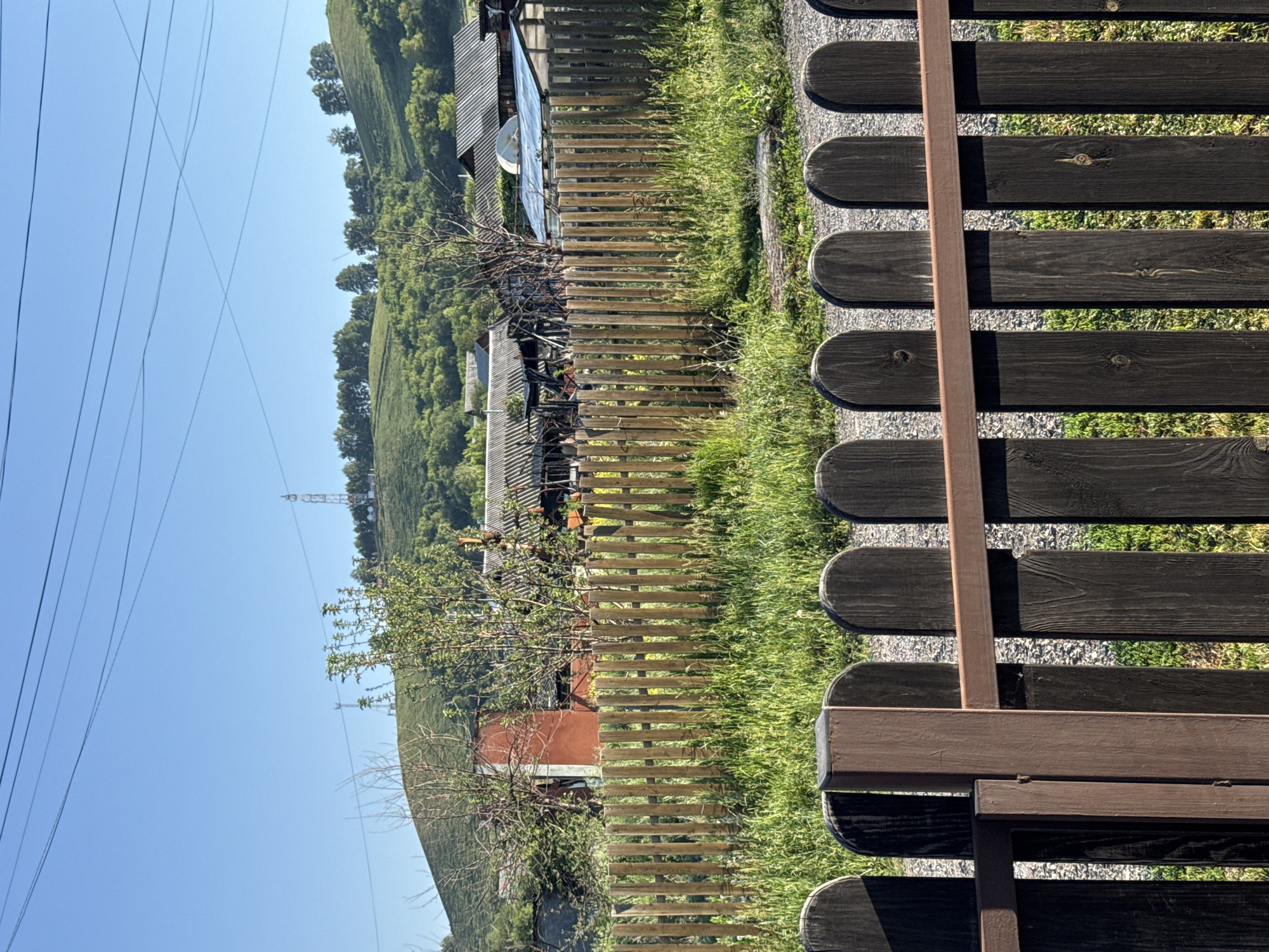 The image shows a rural residential area with houses and fences in the foreground and a green, hilly landscape with trees and communication towers in the background. The sky is clear and blue. In the foreground, there is a dark brown, arched-top picket fence with a metal horizontal bar running across it. Behind this fence is a taller, lighter brown wooden picket fence that is partially obscured by grass. Beyond the fences, several houses with corrugated metal roofs are visible, some appearing in disrepair. There are also trees and vegetation around the houses and on the hill. Two communication towers are visible on the distant hill. The scene is illuminated by natural daylight, casting shadows. There are no people or animals visible in the image. The location is identified as Soloneshnoye, Russia.