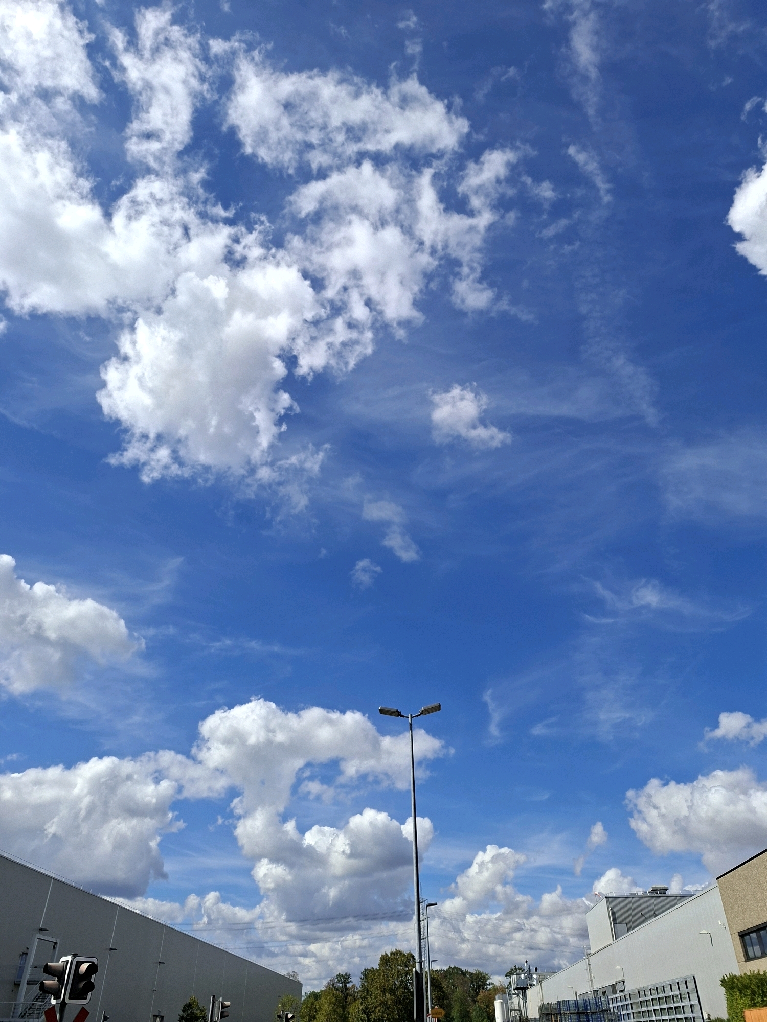 The image captures a bright, daytime scene dominated by a vivid blue sky filled with fluffy white clouds and wispy cirrus formations. In the lower portion of the frame, the edge of a large, light-colored industrial building extends from the left, partially obscuring the view. To its right, a traffic light with red and amber signals is visible. Further to the right, a tall, dark lamppost stands prominently in the foreground. Beyond the lamppost and across the scene, other industrial structures and trees are discernible. The image appears to be taken from an outdoor location in Rastatt, Germany, likely a street or parking area adjacent to an industrial zone.