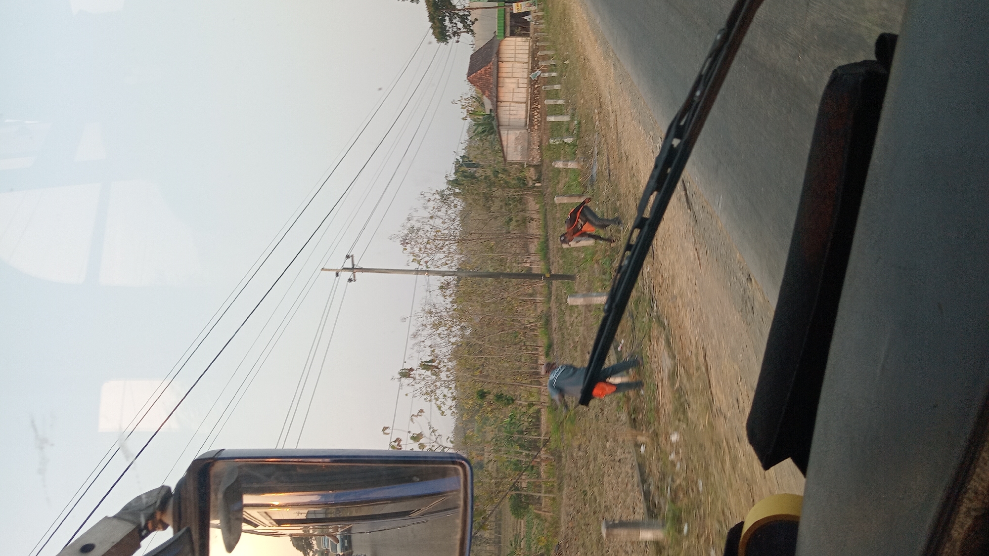 The image, captured from inside a vehicle, looks out onto a rural roadside scene in Klokah, Indonesia. In the foreground, the dashboard and windshield wipers of the vehicle are visible. The scene outside includes a dirt shoulder next to a paved road, with sparse vegetation and trees. Two men are working on the side of the road; one, wearing a gray shirt and orange pants, appears to be raking or sweeping dry leaves, while the other, in an orange and black shirt and gray pants, is also bent over, possibly gathering debris. A utility pole with power lines stands nearby. In the background, there are more trees and a building with a traditional pitched roof. The sky is a pale, hazy blue. The reflection on the vehicle's side mirror shows a road with cars.