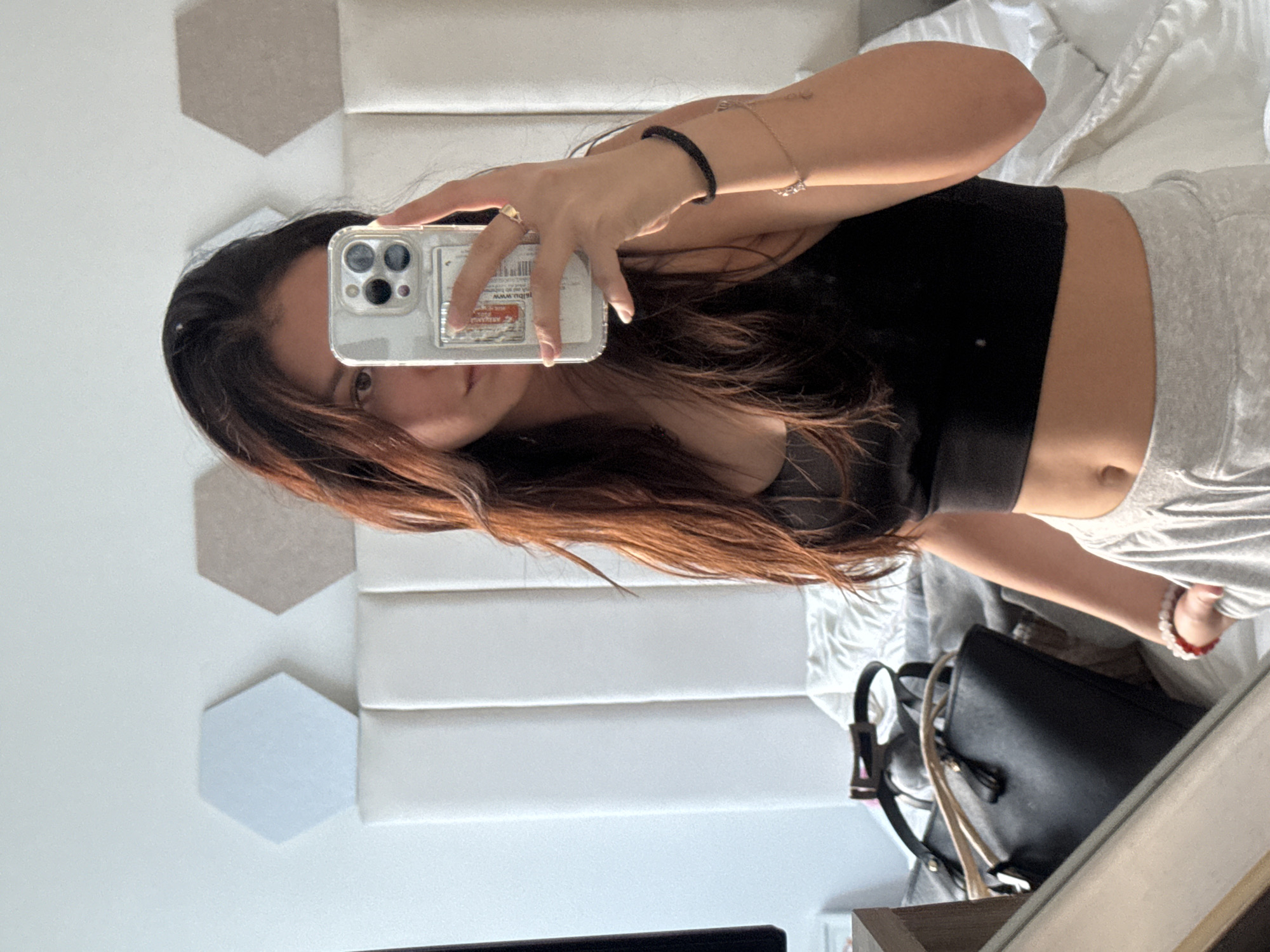 A woman is taking a selfie in front of a mirror. She is holding a smartphone with a clear case. The woman is wearing a black top and gray pants. She has long, brown hair. Behind her, there is a white wall with geometric shapes. A black purse is placed on a surface by the mirror. There is no indication of the location being San Francisco Ocotlán (Ocotlán), Mexico.