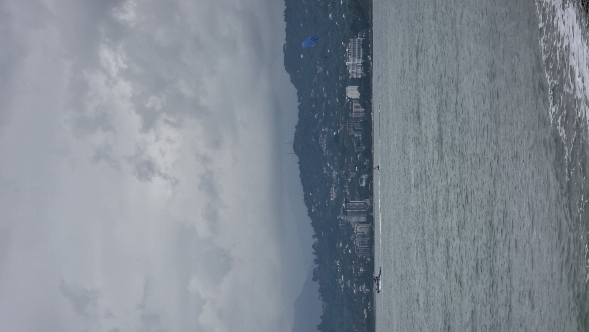 A wide-angle, overcast shot captures the Batumi coastline in Georgia. In the foreground, the choppy, grey sea rolls towards a narrow strip of pebble beach, marked by foamy white waves. The middle ground showcases the bustling city skyline, with a collection of modern high-rise buildings and a sprawling residential area climbing up the lush, green, mist-shrouded mountains that form the backdrop. A small motorboat with two occupants cuts through the water on the left side of the frame. To the right, a blue parachute soars through the cloudy sky, with a person visible below it, suggesting a parasailing activity. The overall atmosphere is subdued, characteristic of a cloudy day.