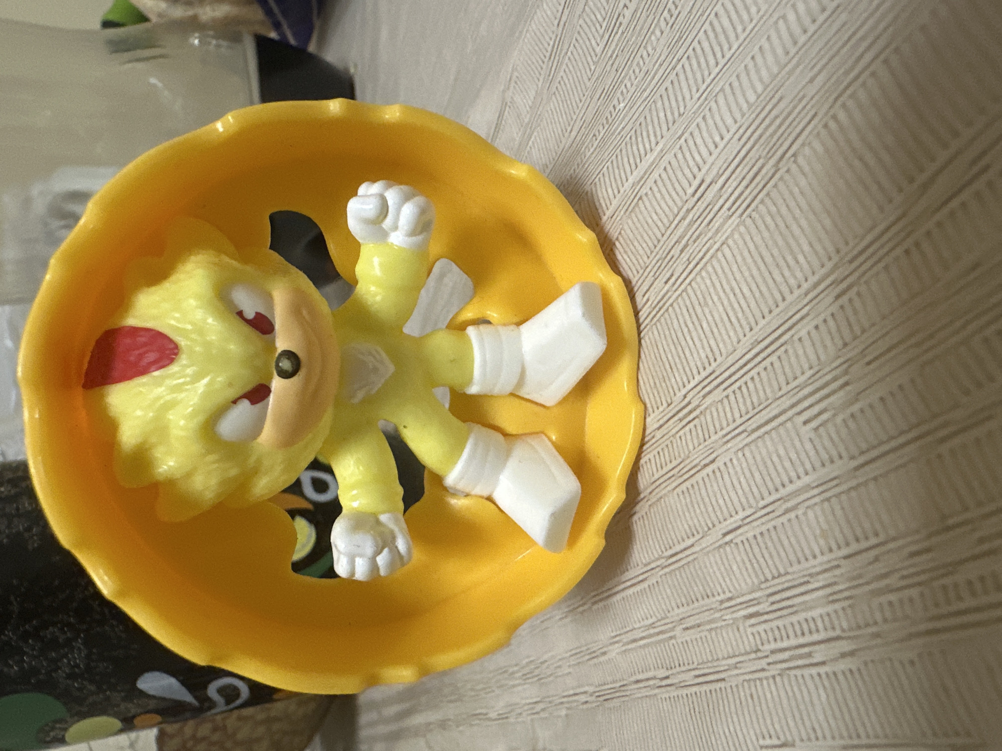A yellow plastic toy figure, resembling Super Sonic from the Sonic the Hedgehog franchise, is the central subject of the image. The figure is bright yellow with white gloves and shoes, spiky quills, and a distinct red marking above its left eye. It lies on its back within a yellow, scallop-edged plastic container, which appears to be a small bowl or promotional item.
The container and toy rest on a flat surface against a light-colored, textured background that exhibits linear patterns, resembling wallpaper or a woven fabric. In the lower-left corner, beneath the yellow container, a blurry dark object with abstract white, green, and yellowish patterns is partially visible. Further in the upper-left background, out-of-focus domestic items and light surfaces can be discerned.
The scene is set indoors, likely in a home in Blagoevgrad, Bulgaria, under ambient or artificial lighting. No specific activity is taking place, and there is no visible text or discernible emotional content.