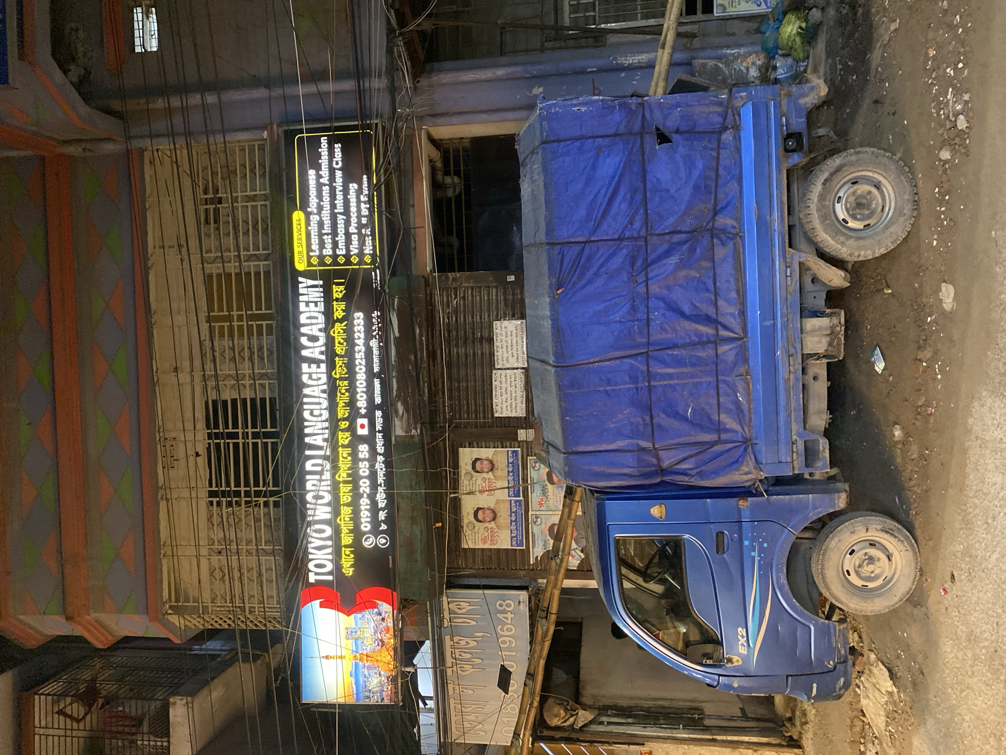 The image depicts a street scene in Dhaka, Bangladesh, at night. A blue truck with a covered cargo bed is parked on the right side of the frame. On the left, a building displays a sign for "TOKYO WORLD LANGUAGE ACADEMY," offering Japanese language learning, visa processing, and other services. The sign includes phone numbers. Posters are visible on the building near the truck. The ground appears littered and uneven. Power lines crisscross overhead.