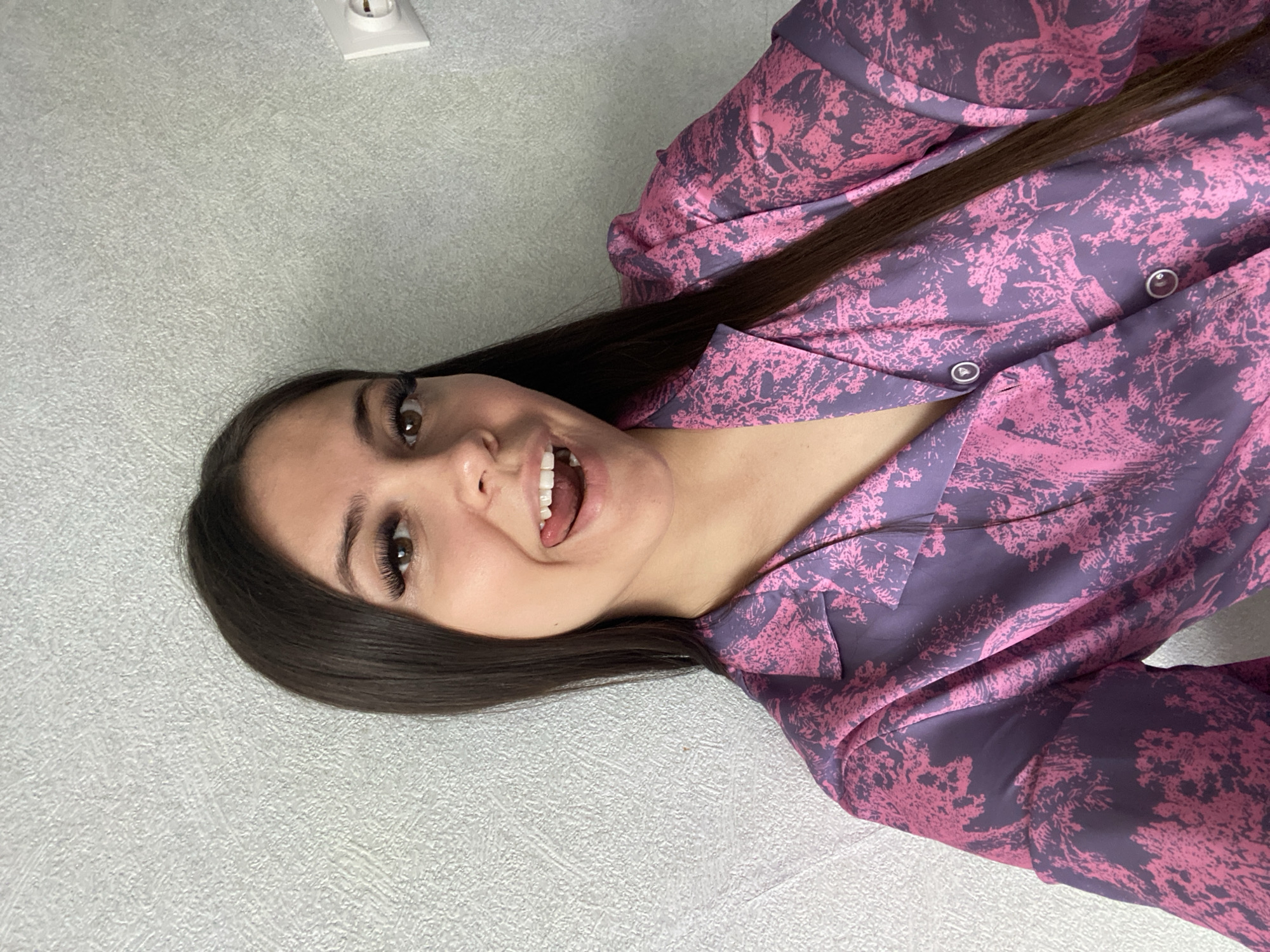 A young woman with dark hair and brown eyes is smiling and sticking her tongue out at the camera. She is wearing a patterned pink and purple button-down shirt. The background is a light grey wall with a white electrical outlet. The image was possibly taken indoors. I am unable to determine if this picture was taken in the city of Chaikovsky, Russia.