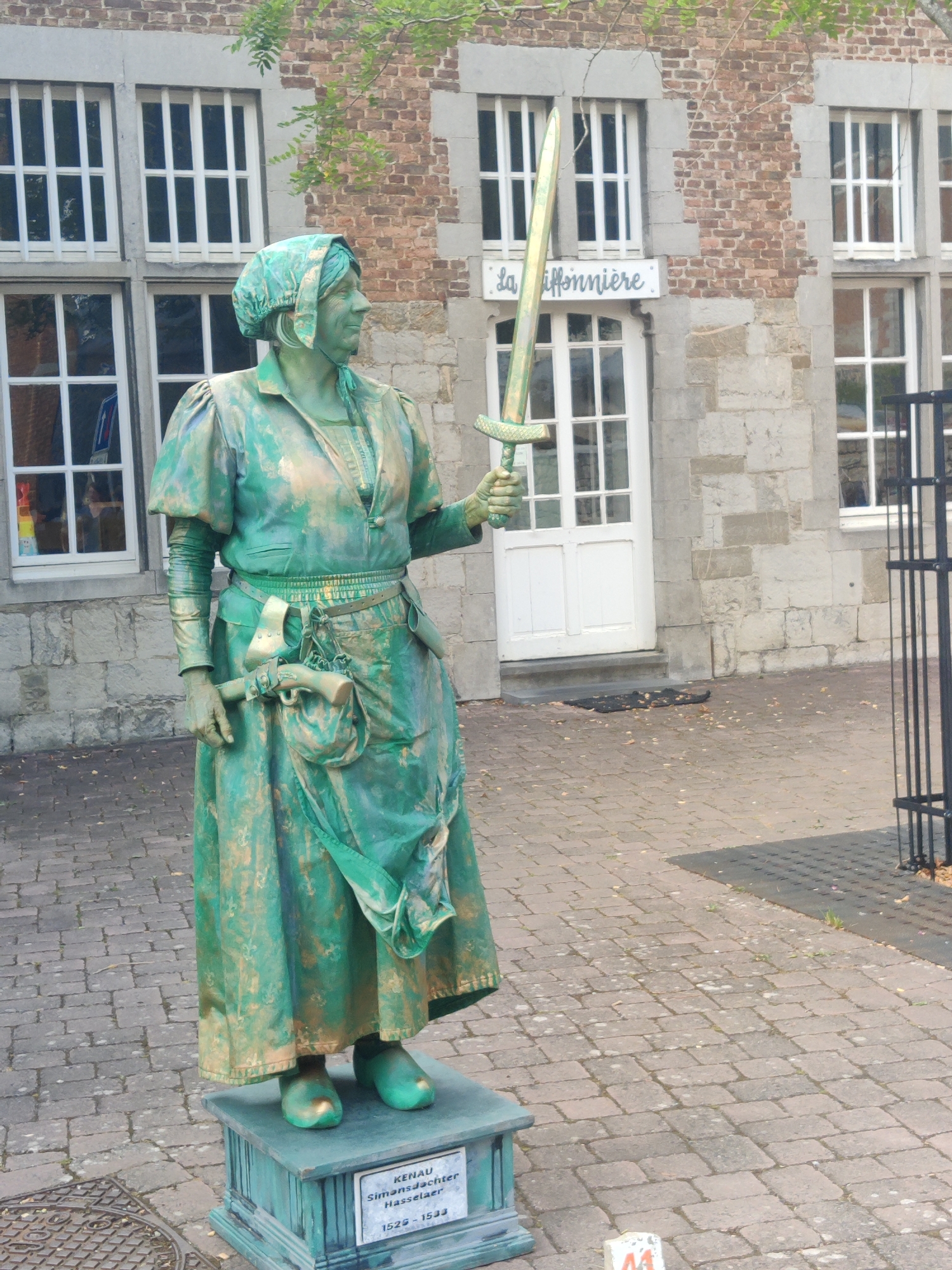 A full-body, three-quarter shot shows a statue of a person painted entirely in green with gold accents. The statue is a woman dressed in historical attire, wearing a headscarf, a long-sleeved blouse with puffed shoulders, a cinched waist with a belt, and a floor-length skirt. She holds a sword in her right hand, raised upwards. A pouch and a pistol are attached to her belt. The statue stands on a teal-colored pedestal with an inscription that reads "KENAU SimonsJachter Hasselaer 1525-1533". The statue is positioned on a cobblestone pathway in front of a building with brick and stone facade, featuring multiple white-framed windows and a white door. A street sign is visible above the door with the text "La Hiffonniere". The overall scene suggests an outdoor historical display or a street performance, likely in a public square or street within Marche-en-Famenne, Belgium, as indicated by the inscription on the pedestal, which refers to Kenau Simonsdochter Hasselaer, a historical figure.