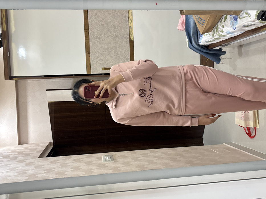 A person in a pink tracksuit takes a mirror selfie. The top of the tracksuit has a small zipper and says "Country Club" with a crest. The pants have a white stripe down the side. The person is holding a maroon-colored smartphone with nail art on their fingers. A bed with items on it can be seen, along with a dresser and patterned wallpaper. The photo is taken at an angle and is inside a mirror frame.