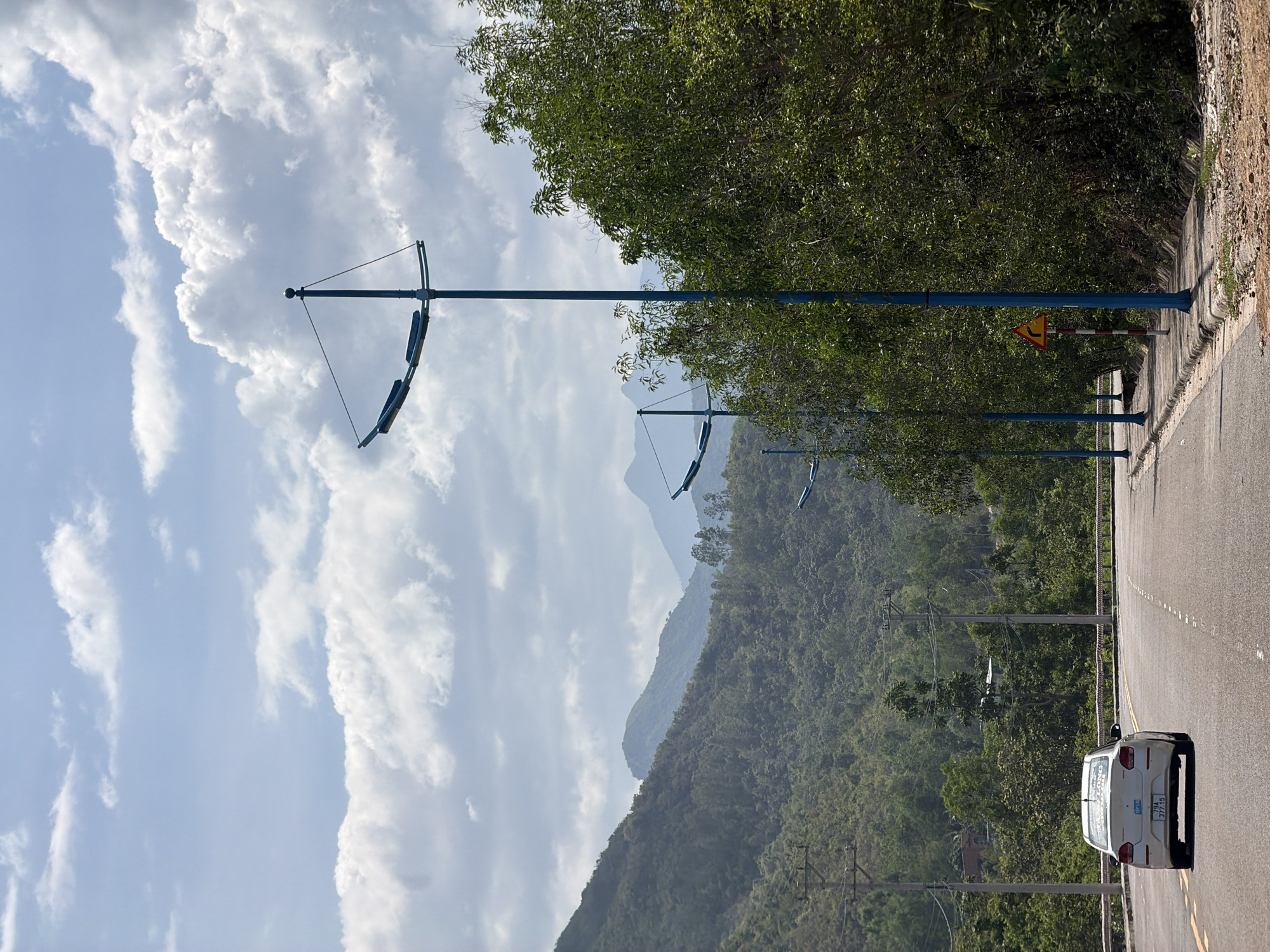 A white car is driving on a paved road with yellow lane markings. The road curves to the left, and on the right side of the road, there is a sidewalk with blue lampposts and a yellow warning sign indicating a sharp curve to the right. A dense green forest covers the hills in the background, and the sky is filled with white and gray clouds. The scene appears to be on the outskirts of Vĩnh Lương, Vietnam, possibly along a scenic route.
