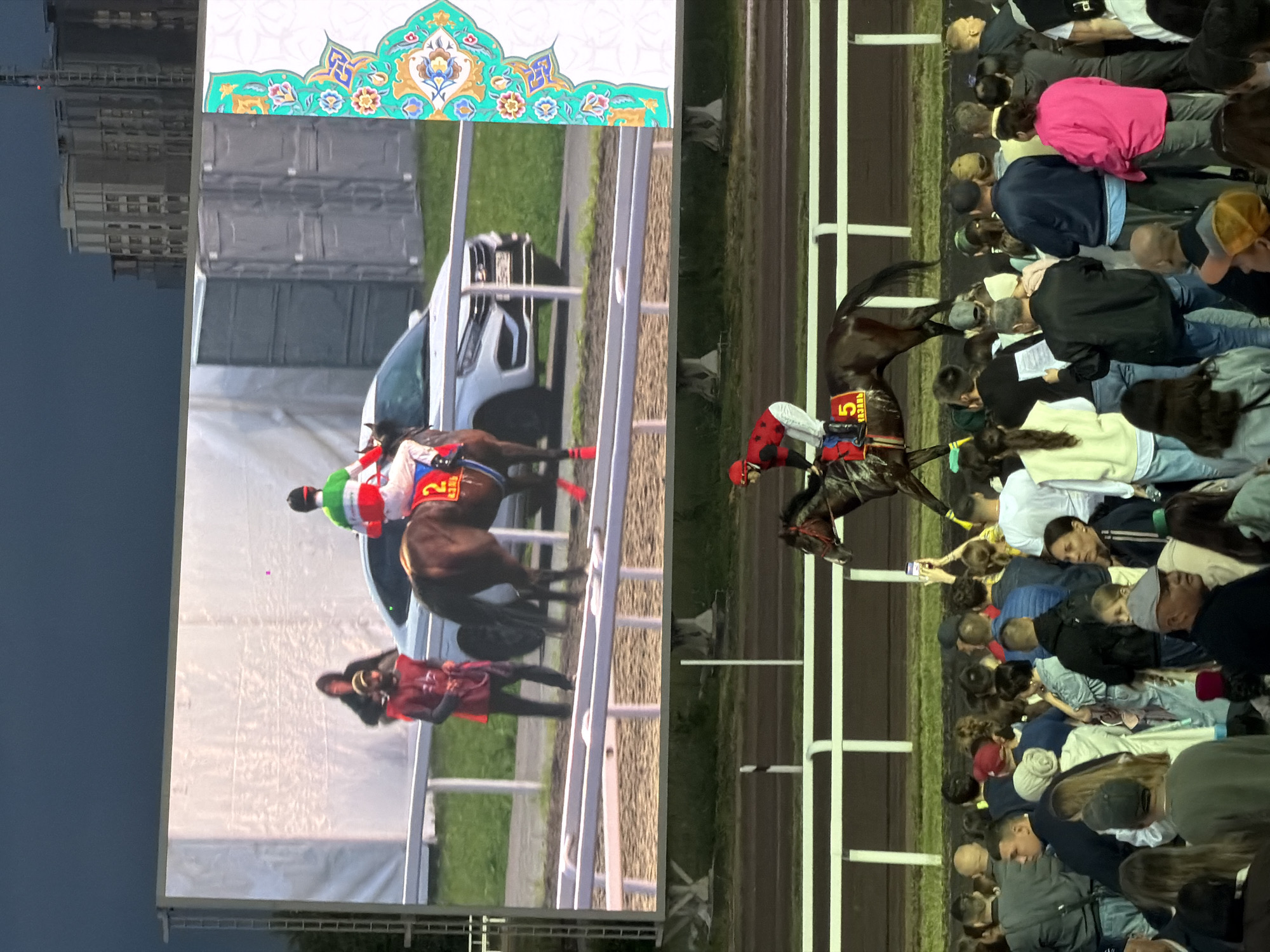 The image depicts a horse race being shown on a large outdoor screen, with a live race occurring in the foreground. In the screen's display, a jockey in green, white, and red silks rides a brown horse with the number "2" on its saddlecloth. A white car and two portable toilets are visible behind the screen's track. To the right of the screen, an ornate, colorful pattern with floral motifs is displayed. In the foreground, a jockey wearing a red and black uniform with the number "5" on their saddlecloth rides a brown horse on a dirt track. A crowd of spectators is gathered behind a white fence, watching the live race. The scene appears to be taking place in the evening, with the sky dark and buildings visible in the distance.