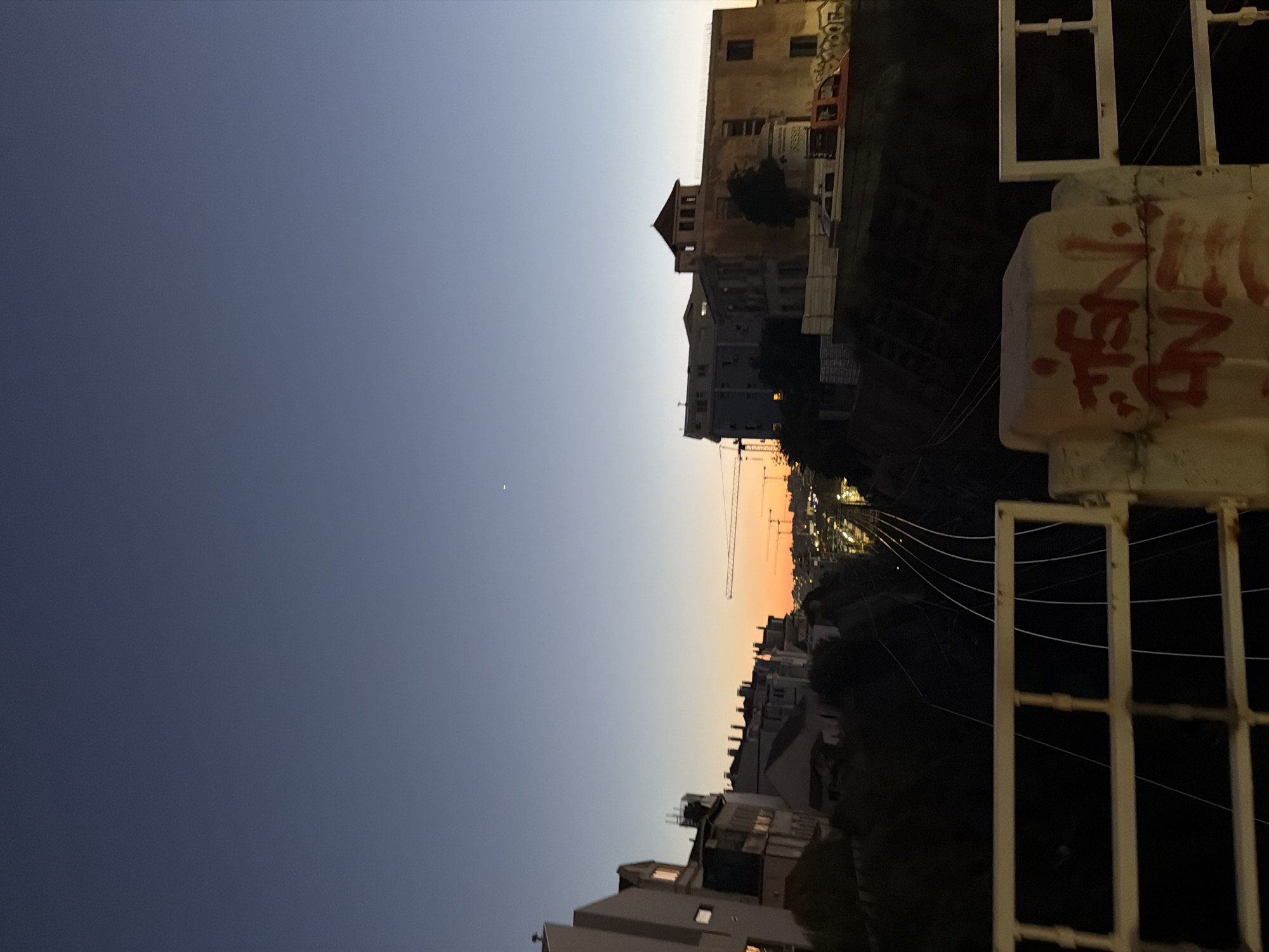 The image captures a twilight scene in Cascais, Portugal, likely overlooking a railway line. In the foreground, a white railing, partly obscured by graffiti, borders the viewpoint. Beyond the railing, multiple railway tracks recede into the distance, flanked by dark, sloped embankments. Along the crest of the embankments, buildings with various architectural styles are visible, some featuring distinct rooflines and windows. In the mid-ground, construction cranes are present, suggesting ongoing development or infrastructure work. The sky transitions from a deep blue at the top to a vibrant orange near the horizon, indicating the sun has recently set. A single, distant star or planet is visible in the upper right quadrant of the sky. The overall atmosphere is calm and urban, with hints of industrial activity due to the railway and cranes.