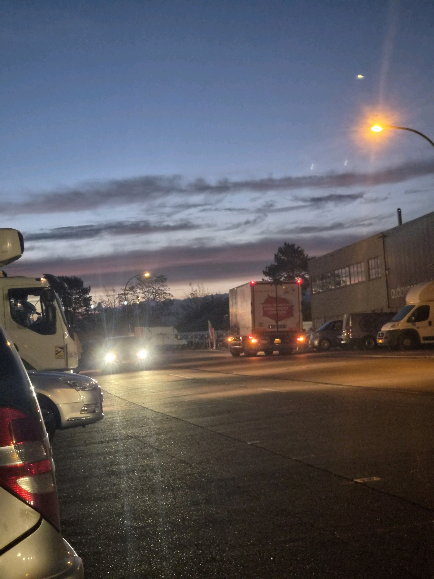 The image depicts a twilight scene on a wet asphalt surface, likely a road or parking lot, in or near Leudelange, Luxembourg. The sky is a gradient of deep blue to dusky purple, with striated clouds. A street lamp with a bright orange glow illuminates the upper right portion of the frame. In the foreground, the rear of a silver car is visible on the left. Moving towards the right, several vehicles are present, including a white truck with its hazard lights illuminated, a white panel van, and what appears to be a white truck with a refrigeration unit on top. Further to the right, a large industrial-style building with multiple windows is visible. The overall atmosphere suggests a transition from day to night, possibly in an industrial or logistical area.