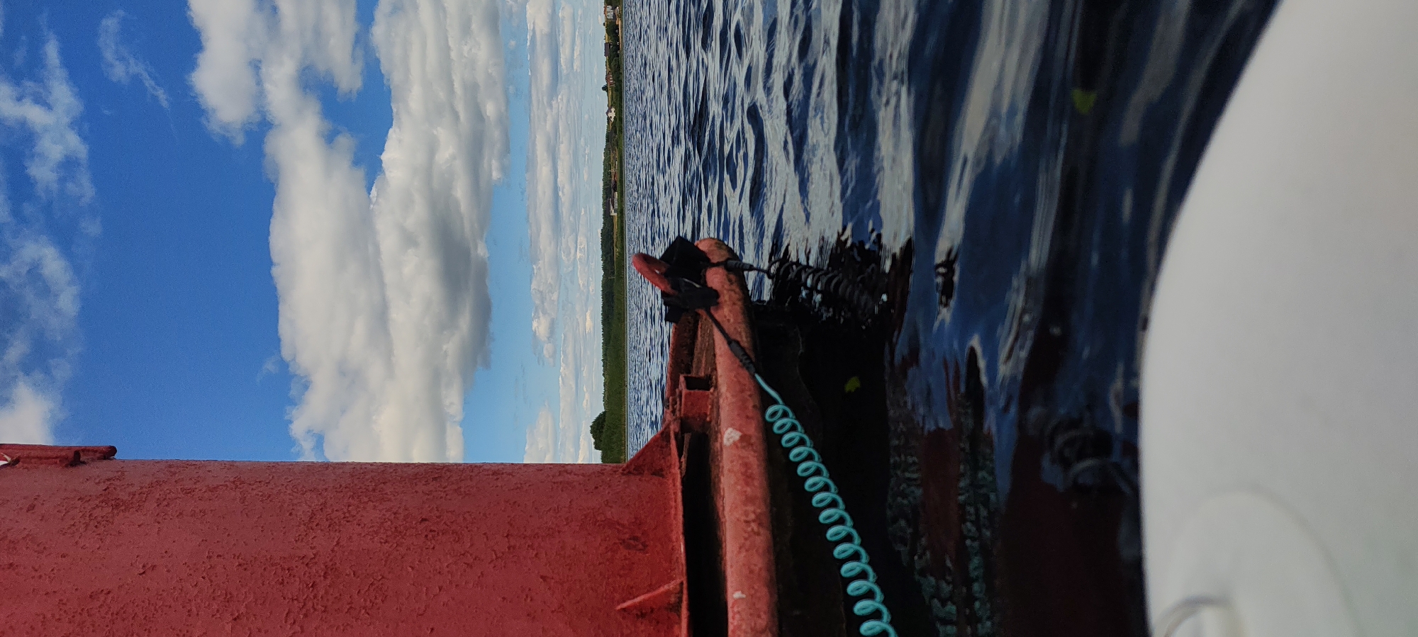 The image shows a close-up view of a red buoy floating on dark blue water. A coiled turquoise rope is attached to the buoy and extends into the water. In the background, there is a clear blue sky with white clouds, a line of green trees, and several small houses in the distance. The location is identified as Щиглицы, Russia. The scene appears to be a calm day on a body of water, possibly a lake or a river, with a buoy marking a navigation channel.