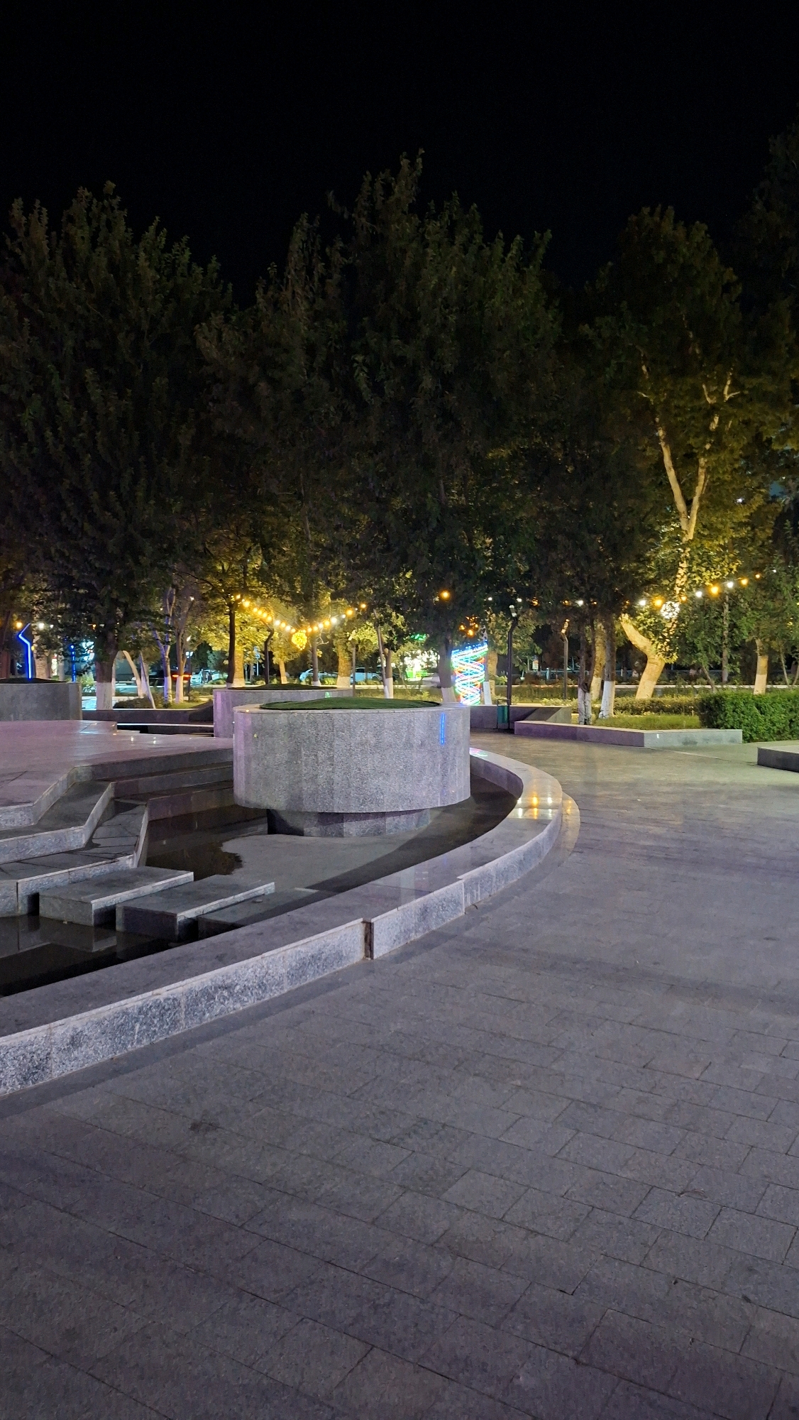 An exterior, night-time, vertical, eye-level, medium shot captures a public park area in Navoiy City, Uzbekistan. In the foreground, a grey, stone-tiled walkway curves to the right, leading the eye towards a multi-tiered water feature. This water feature consists of grey stone ledges and a central, circular, stone structure. Trees with dark green foliage line the background, illuminated by strings of yellow lights draped between them. The night sky is dark, with no visible celestial bodies. The scene is devoid of people.