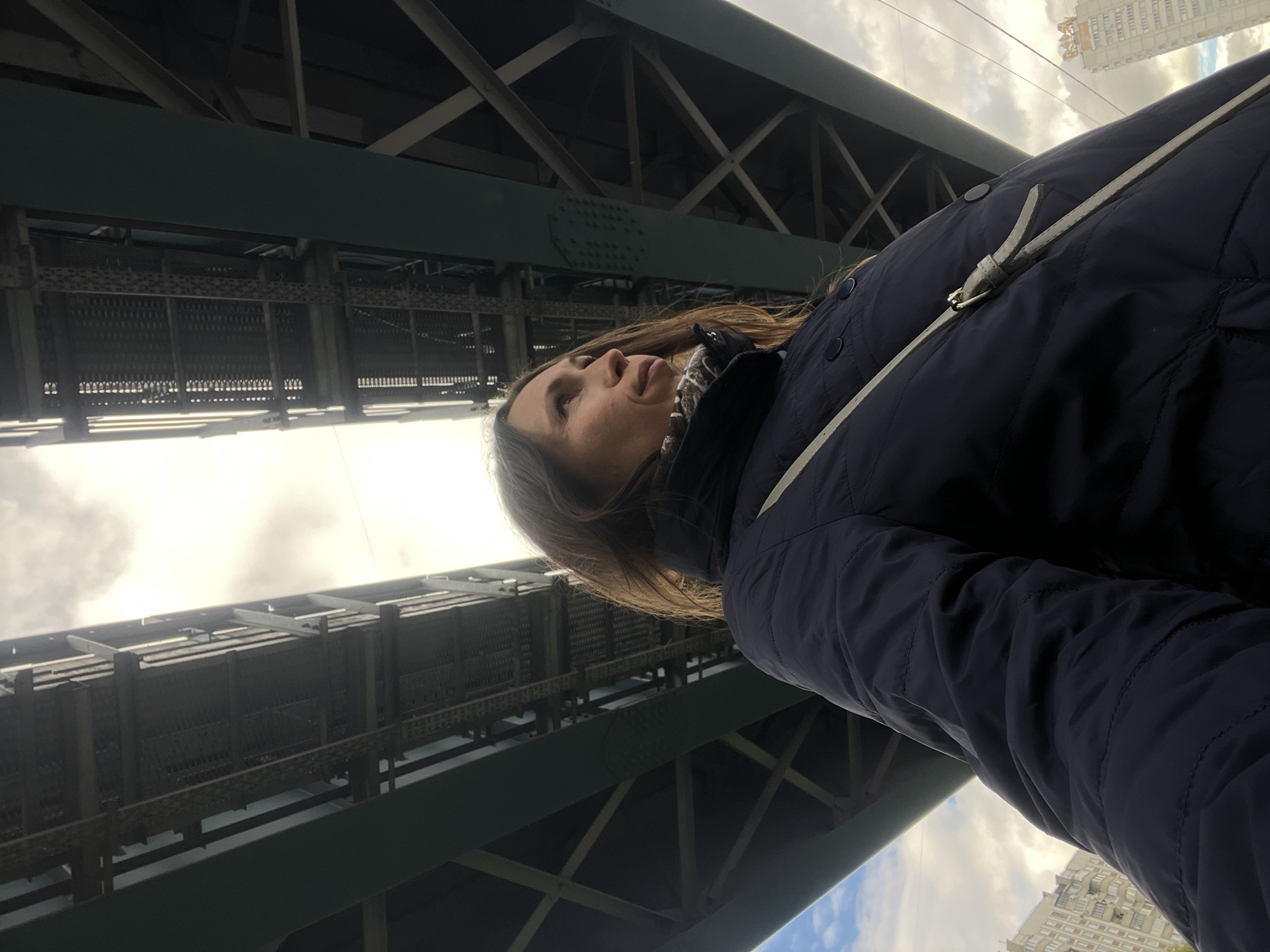 A low-angle shot looking up shows a woman standing underneath a large bridge structure. The bridge appears to be made of green-painted metal with an intricate framework. A building is visible in the background. The woman has fair skin and brown hair and is wearing a dark blue quilted jacket with a white strap across her torso. The location is likely the Moscow Metro Bridge in Russia.