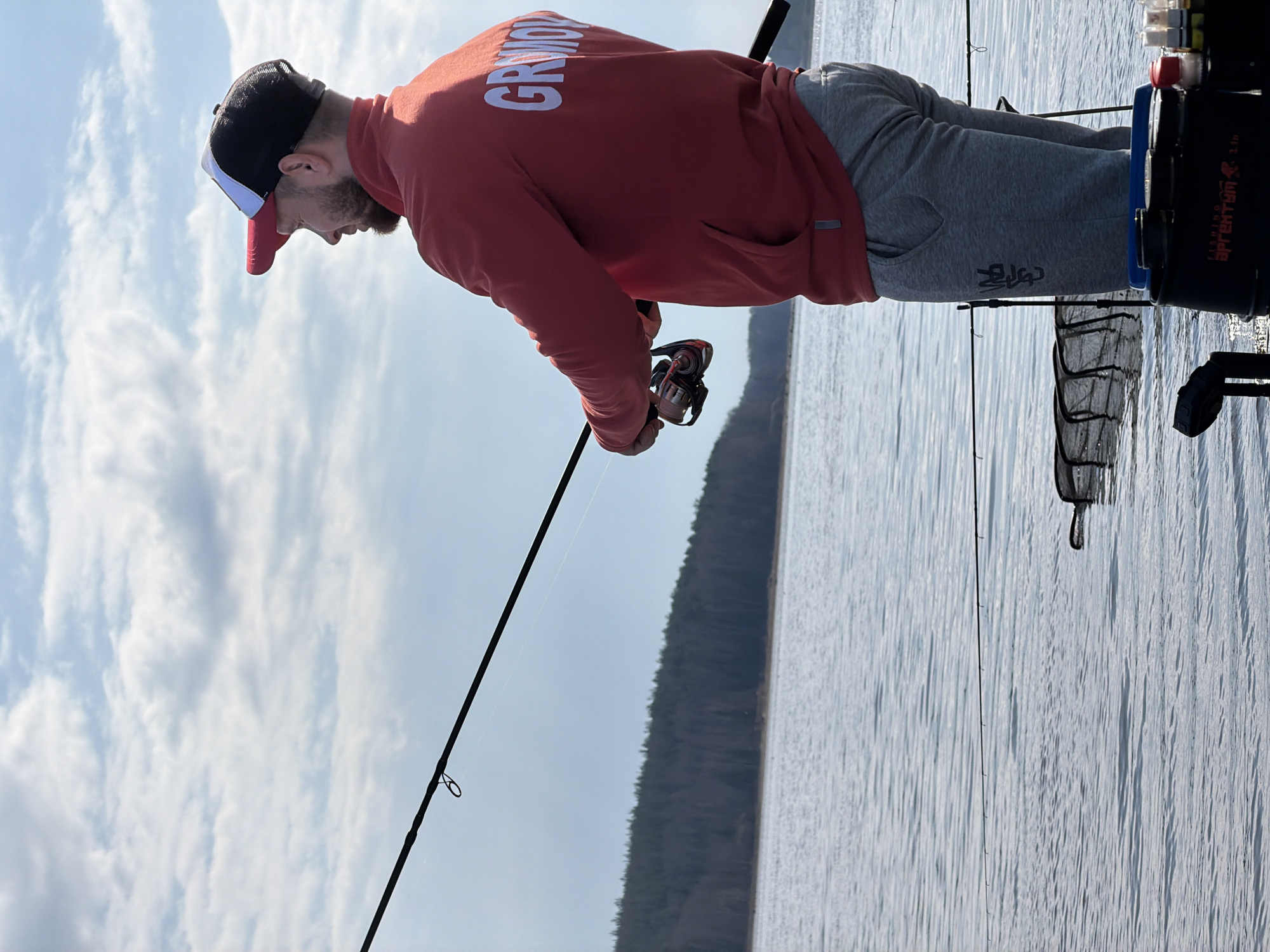 A man is fishing on a body of water. The man is wearing a red hooded sweatshirt with white lettering, gray sweatpants, and a baseball cap. He is holding a fishing rod and reel. A tackle box and net are behind him. The location is likely in the city of Chaikovsky, Russia, given the tag associated with the image. The water reflects the sky, which is partly cloudy. A forested shoreline is visible in the background.