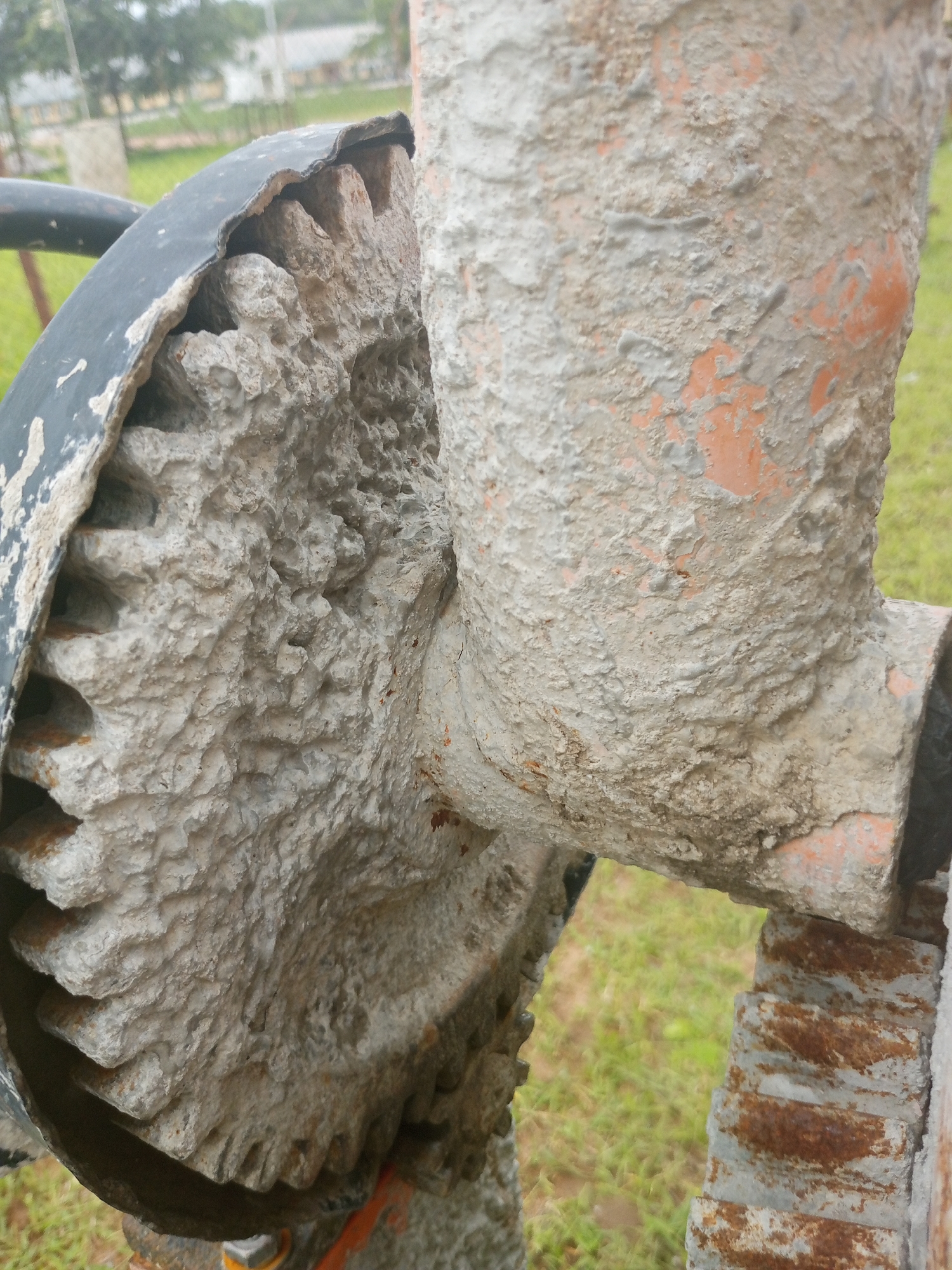 A close-up shot of a cement mixer's gear and support structure, heavily coated in dried cement. The gear, made of metal with visible teeth, is partially obscured by the thick, gray cement. The supporting pipe structure, painted orange and also caked in cement, extends upwards and to the right. The background is out of focus, showing green grass and some buildings in the distance. The scene suggests a construction or industrial setting, likely outdoors in Jalingo, Nigeria, given the visual cues of the environment.