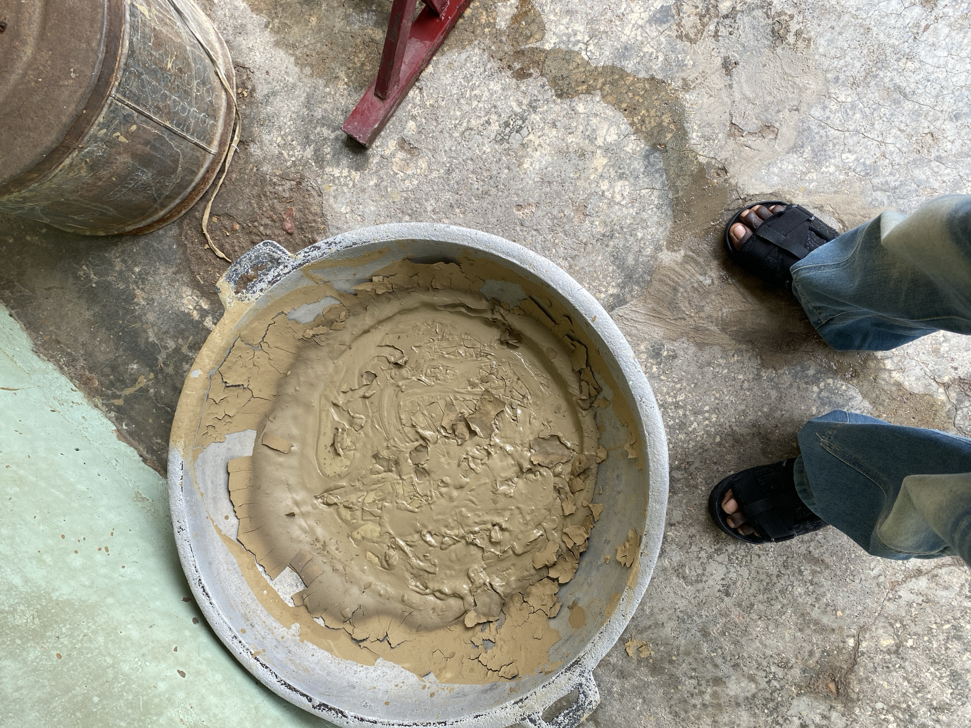 The image depicts a scene outdoors, likely in Abeokuta, Nigeria, characterized by a rough, concrete floor with patches of dirt and water. In the center, a large, weathered metal basin is filled with a mixture of mud and water, showing signs of cracking and drying around the edges. To the right of the basin, a red, wedge-shaped object, possibly a shim or lever, rests on the ground. Further to the right and slightly behind the basin is a tall, cylindrical metal container, showing significant rust and wear. In the foreground, a person's legs are visible, wearing light blue jeans and black sandals, suggesting their presence near the basin. The background features a light green, textured wall with brown splotches.