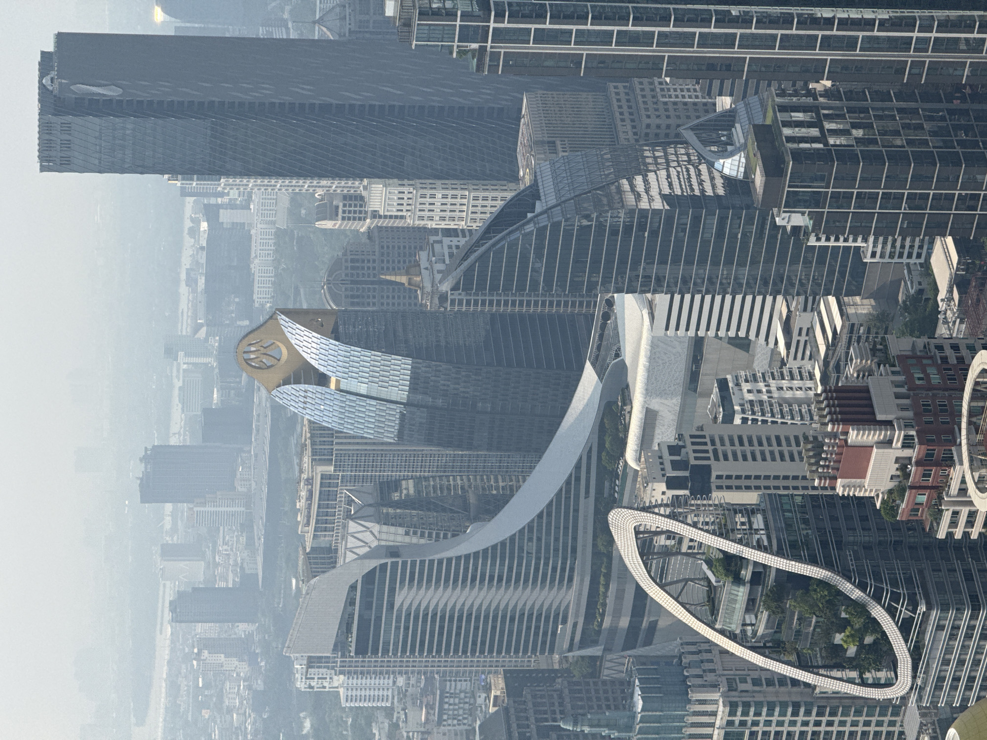 The image shows an aerial view of the city of Bangkok, Thailand. Modern skyscrapers and high-rise buildings dominate the skyline. The Mahanakhon building stands out due to its unique design. In the foreground is the shape of a ring that is part of a building, with trees growing on it. The overall view is somewhat obscured by haze or smog.