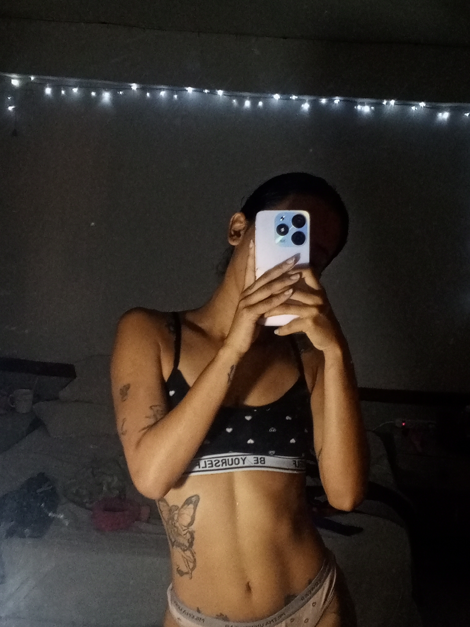 A person is taking a selfie in a room with low lighting. The person is wearing a sports bra with a heart pattern and underwear with a heart pattern, exposing their abdomen, which has a butterfly tattoo. The person is holding a smartphone with multiple camera lenses in front of their face. Behind the person, there is a bed with bedding and a string of lights hanging on the wall. The location is not discernable as being in Porlamar, Venezuela.