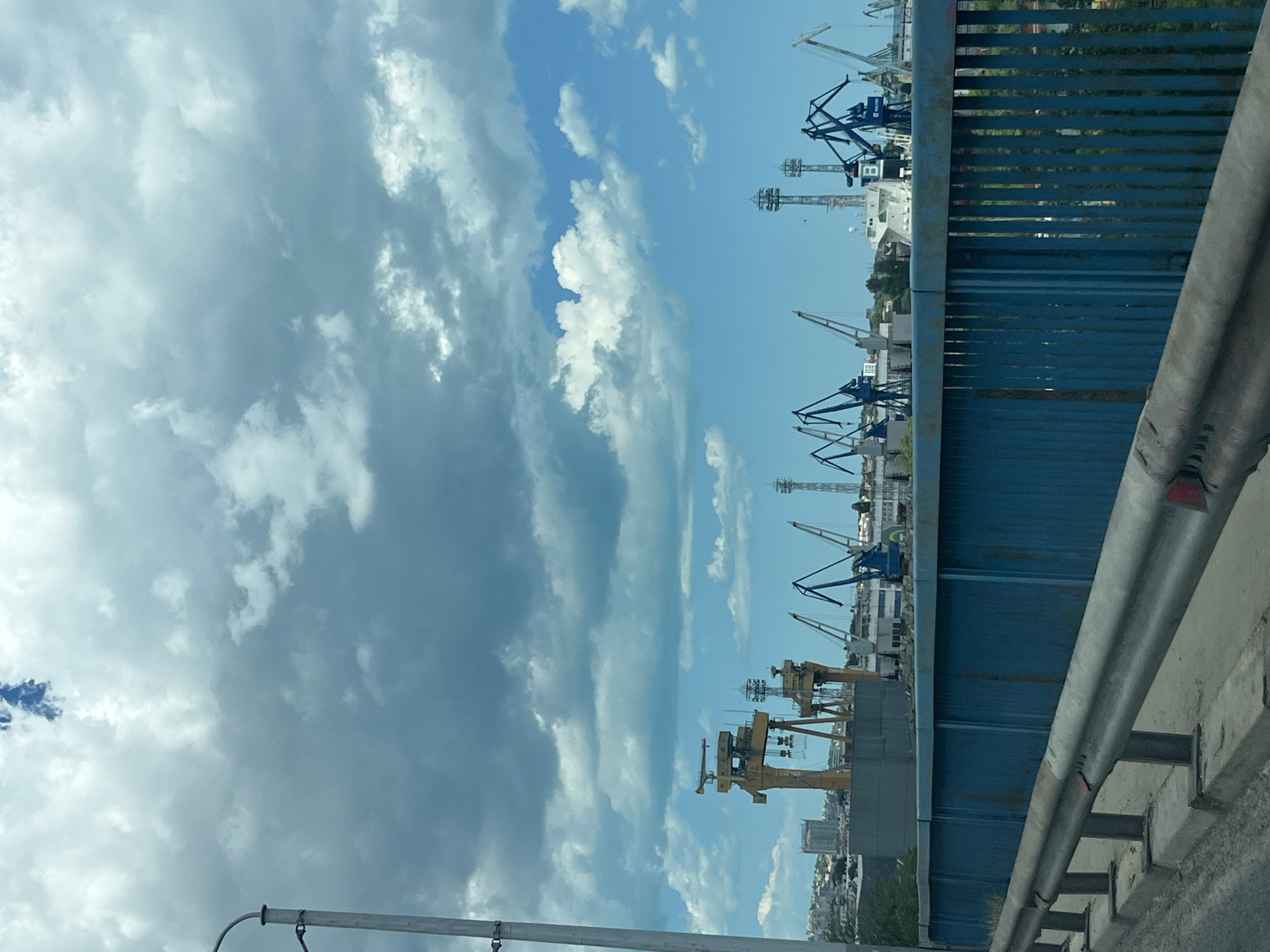 This image depicts a wide view of a port area, likely in the city of Varna, Bulgaria. In the foreground, a blue metal fence and a concrete barrier with a metallic railing are visible, suggesting the vantage point is from a bridge or elevated walkway. The background is dominated by a dramatic sky with a mix of dark, heavy clouds and bright, blue patches. Below the sky, a cityscape with buildings, including a tall, slender tower, can be seen on the left. The central focus of the port area is a collection of large industrial cranes of various designs and colors, including several prominent yellow gantry cranes. Smaller blue cranes are also visible, along with what appears to be a ship or large vessel docked in the harbor. The scene suggests an active industrial or shipping hub.
