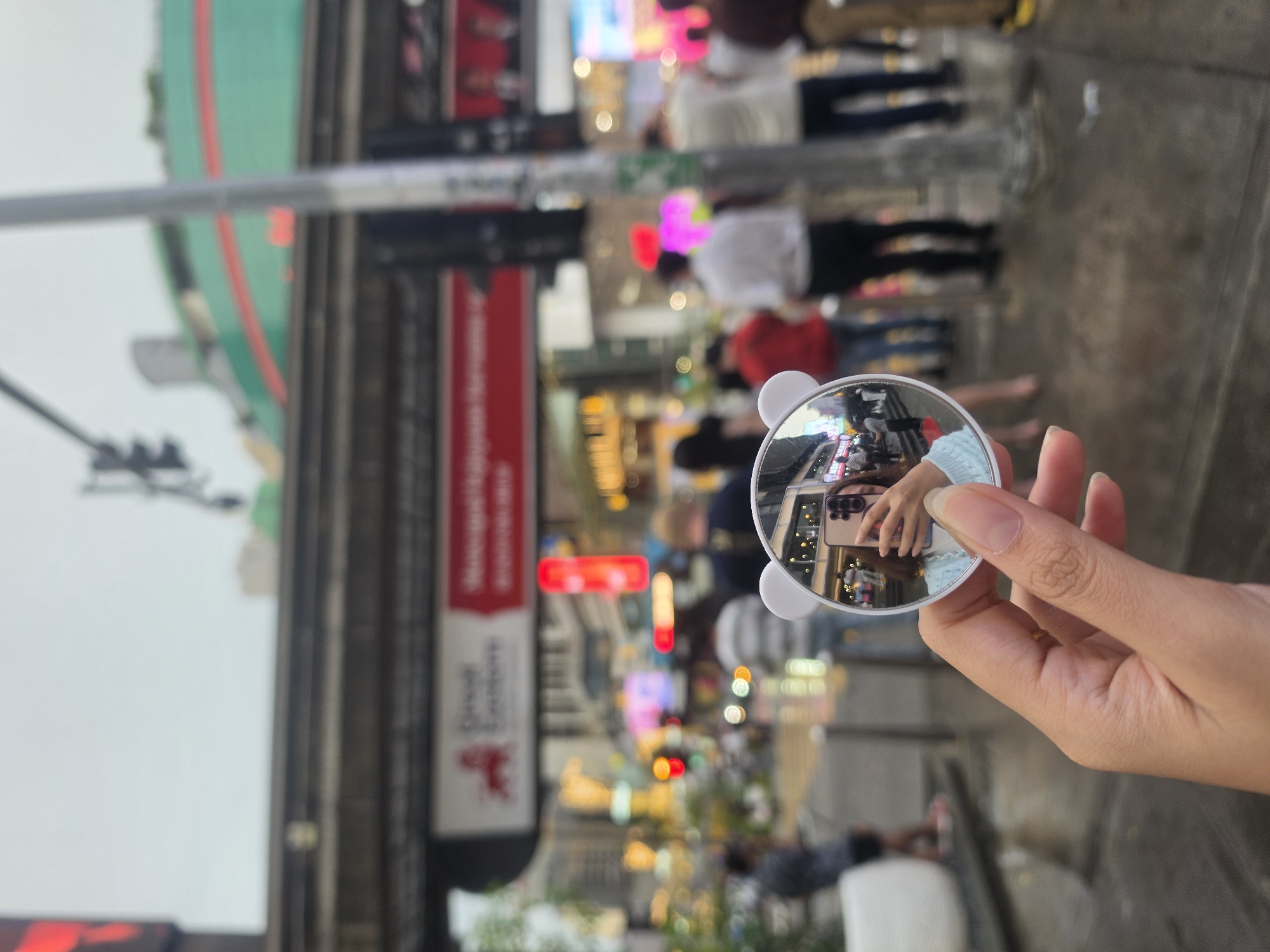 A vertical, eye-level outdoor shot taken in Kuala Lumpur, Malaysia. The image is shot through a small, circular mirror with bear ear-like extensions, held by a human hand. The mirror reflects a person taking a photo of themselves with a smartphone.
The background shows a busy street scene with pedestrians, red traffic lights, and various commercial buildings. The architecture includes modern high-rises with colorful neon signs. A prominent red-and-white sign is visible.
The foreground features the person's hand holding the mirror. The hand and mirror are in focus, while the background is intentionally blurred to create depth of field.
