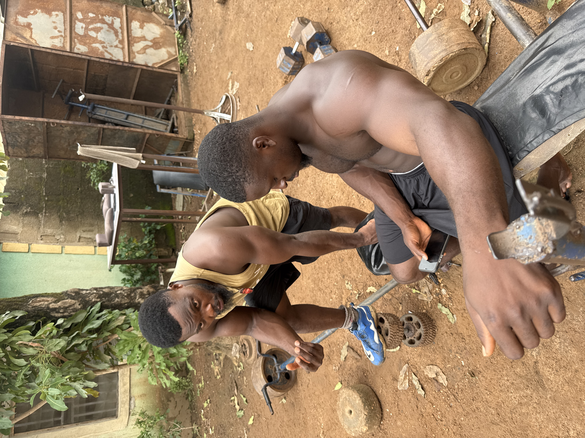 Two muscular Black men are in an outdoor gym setting, likely in Akwanga, Nigeria. The man on the left is standing, wearing a yellow tank top and black shorts with blue sneakers. He is looking at the camera with a serious expression, holding a hand out towards the man on the right. The man on the right is sitting on a black padded bench, wearing black shorts. He is looking down at a smartphone he is holding in his hands. The ground is covered in reddish-brown dirt with scattered dry leaves. In the background, there are various pieces of weightlifting equipment, including old, rustic-looking dumbbells, weight plates that appear to be made of concrete or stone, and a metal rack structure. A tree with green foliage is visible behind the standing man. The overall environment suggests a functional but unpolished outdoor gym.