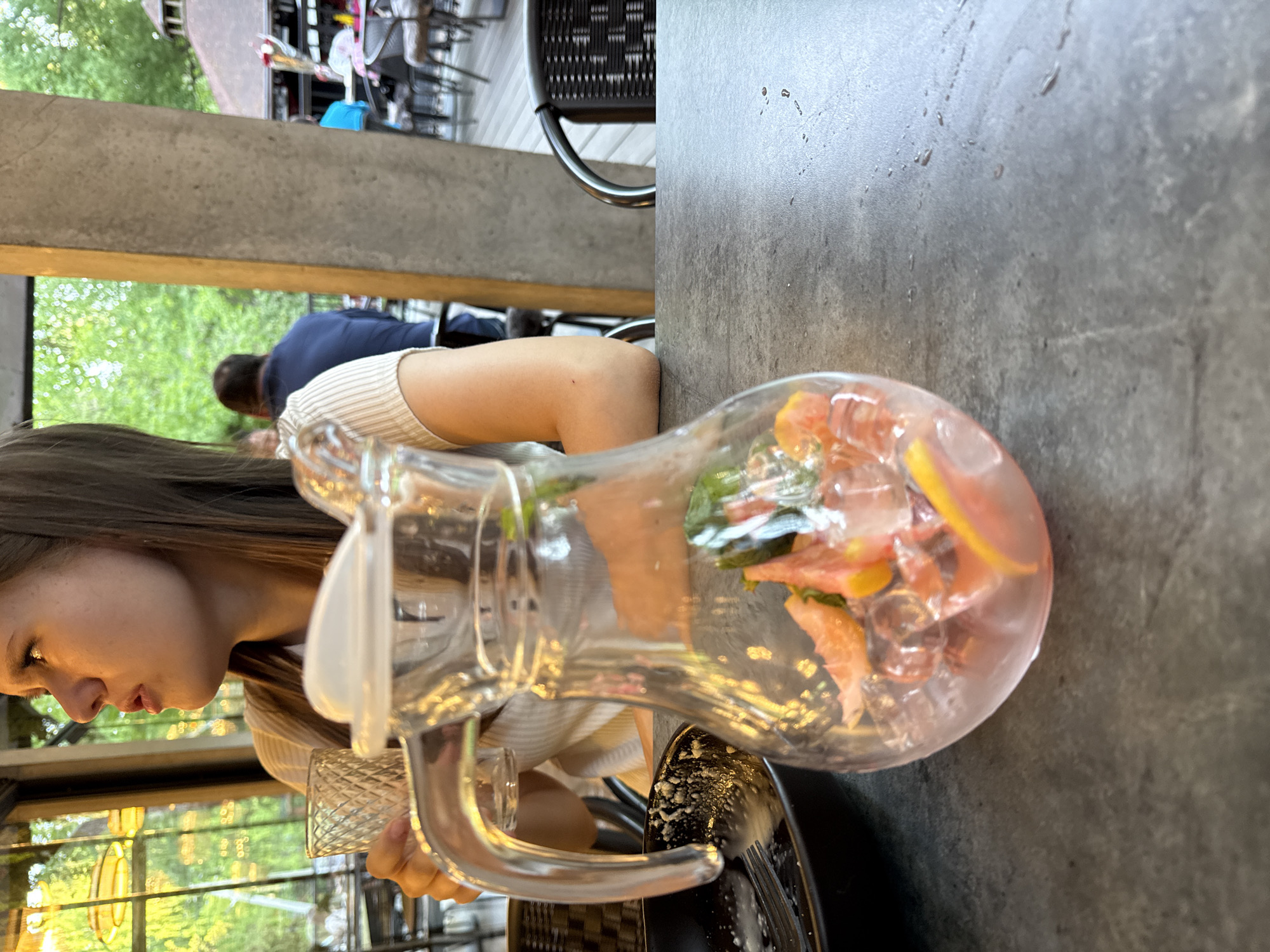 A woman sits at an outdoor table with a pitcher of iced fruit-infused water. The table is grey and rectangular. A concrete support beam is in the background along with greenery and blurred figures at other tables. The location is Izhevsk, Russia.