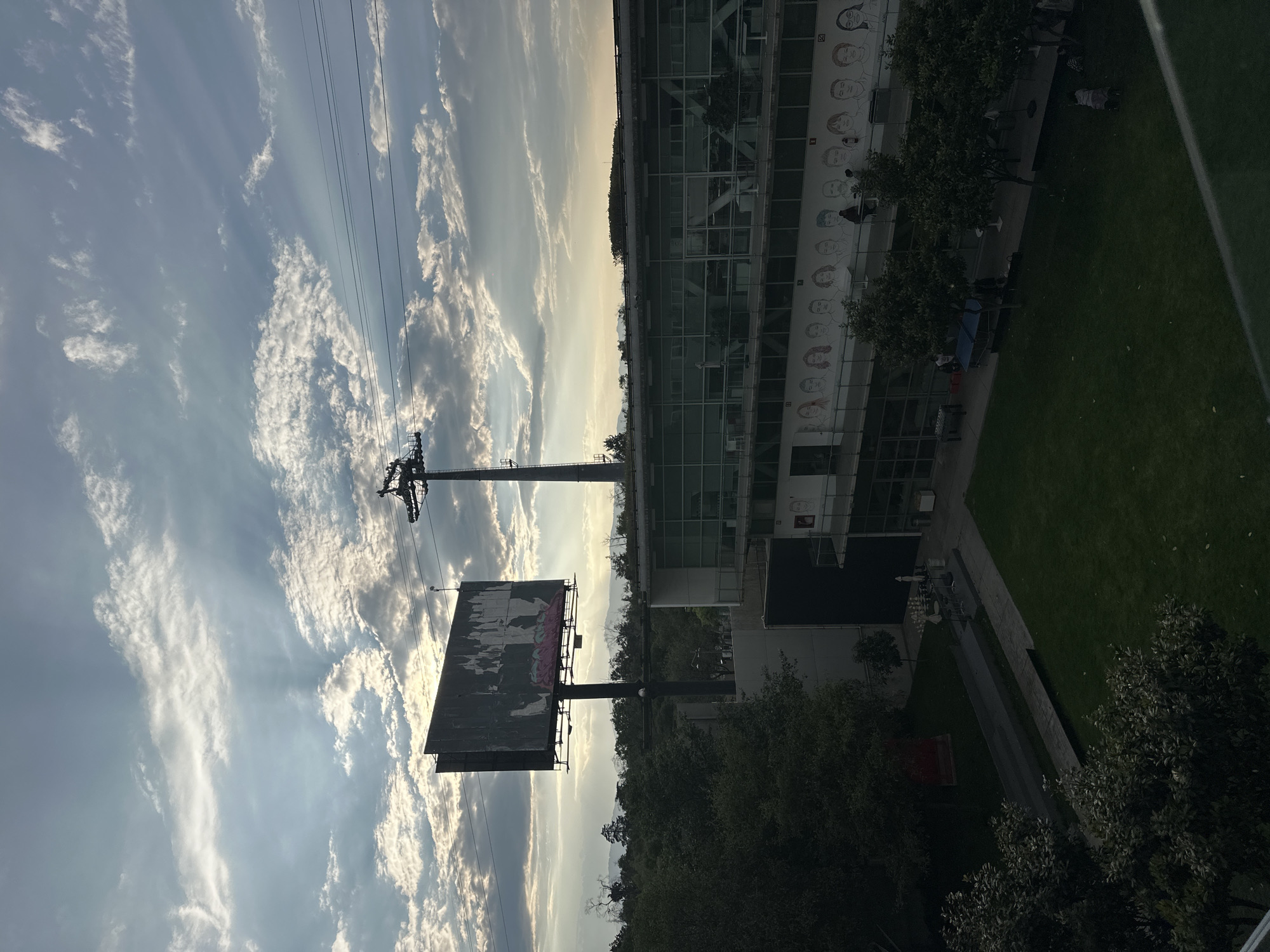The image depicts a view in Mexico City, Mexico, showing an urban landscape with a mix of infrastructure, nature, and architectural elements. The sky is predominantly light blue with scattered white clouds. An electrical power transmission tower and overhead power lines are visible. A large billboard stands prominently, featuring a faded advertisement. Adjacent to the billboard is a modern, multi-story building with a glass facade; the building has a mural on its exterior. Below, trees are visible. A green lawn and walkways are present. People can be seen near the base of the building.