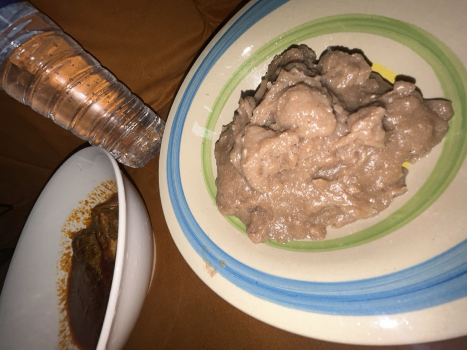 A meal is presented with a plate of brown, porridge-like food, possibly a starchy staple. A separate white bowl contains pieces of meat stewed in a dark red sauce. A plastic water bottle sits nearby. The plates rest on a surface with a warm, brown tone, likely a table or couch. The scene is brightly lit.