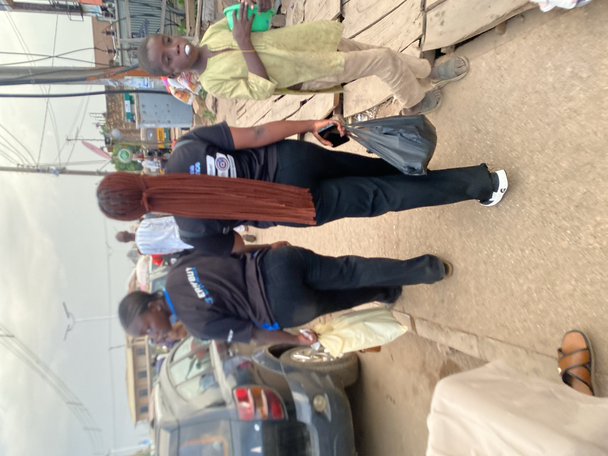 The image depicts an outdoor scene in Bukuru, Nigeria. Two women in dark clothing are observed, with one holding a mobile phone and a black plastic bag. A child with a green container is walking nearby. A car is present in the scene. The environment appears to be a street with buildings and utility poles visible in the background.