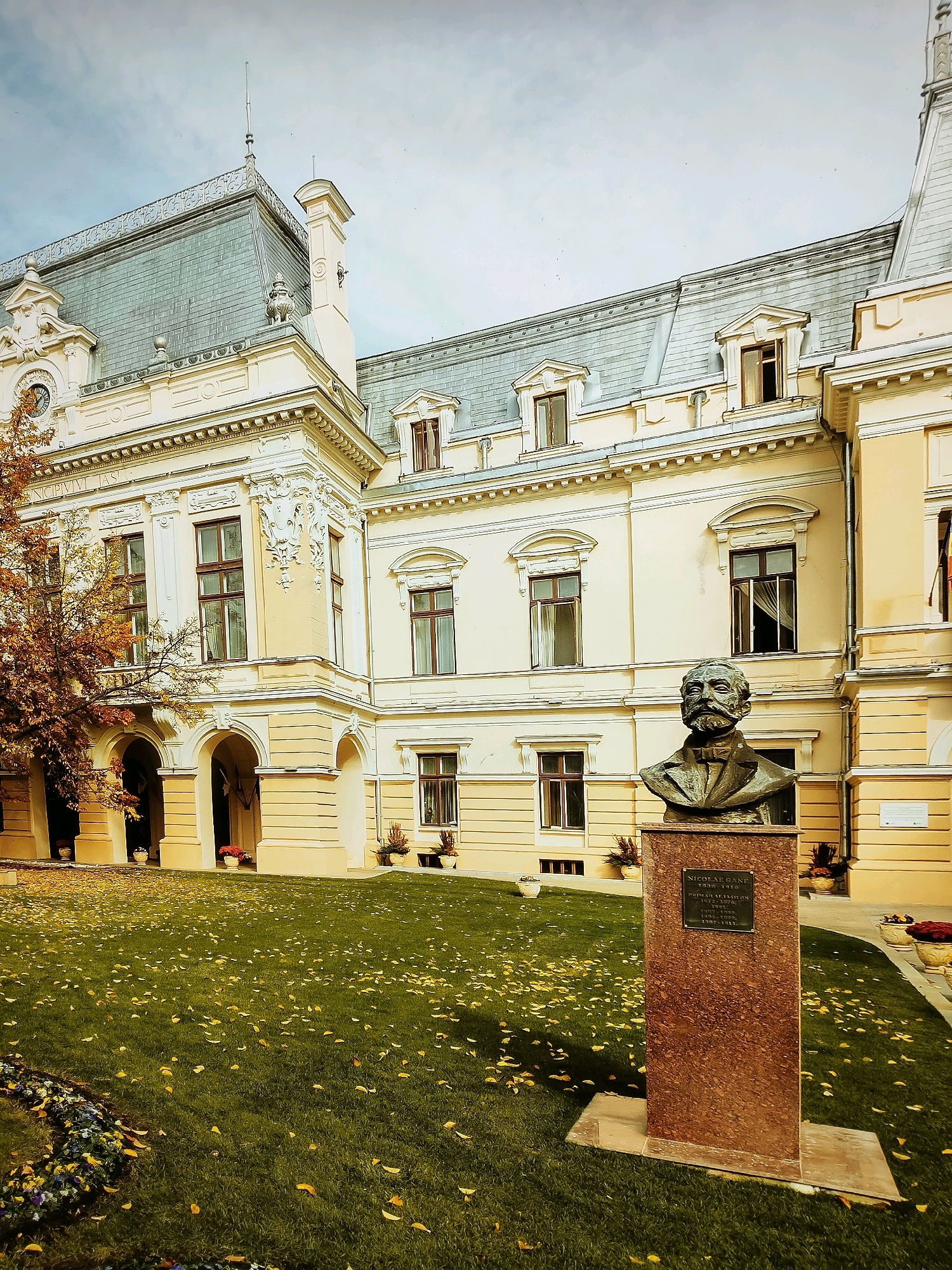 A bronze bust of a man with a mustache and beard sits on a red granite pedestal in front of a cream-colored building. The bust is on the right side of the image, and the building occupies the background. The building has a gray, mansard roof with dormer windows. The facade is adorned with decorative moldings and has several windows with dark frames. A tree with yellow and orange leaves is on the left side of the image, partially obscuring an arched entryway of the building. The ground in front of the building is covered in green grass with scattered yellow leaves. The sky is overcast. The pedestal has a plaque with text, but it is illegible.