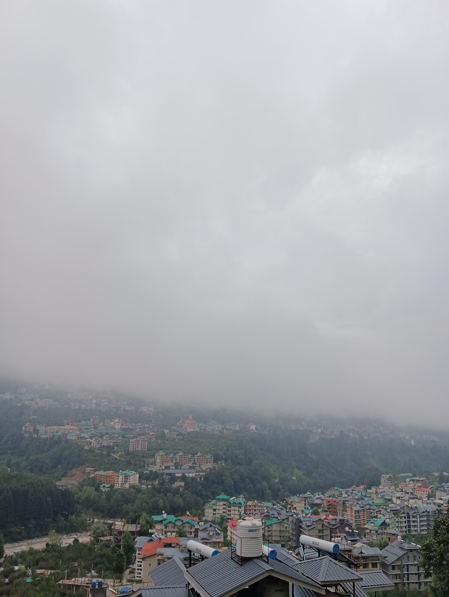 This is a wide-angle, eye-level, outdoor, daytime photograph taken in the city of Chhyal, India. The image captures a hazy, overcast day with heavy clouds obscuring the upper half of the frame and rolling down the distant mountainside. In the foreground, there are several buildings with grey, sloped roofs, some of which are equipped with solar water heaters and tanks. These buildings appear to be residential or hotels, with multiple stories and visible windows.
Further down the hillside, a dense cluster of buildings with various colored roofs, predominantly green and red, stretches across the landscape. These buildings are closely packed together, suggesting a town or city center. The architecture includes multi-story structures with balconies and numerous windows. The middle ground is dominated by lush green trees and the densely packed urban development.
The background features a mountainous terrain, with the city's buildings climbing the slopes. The higher parts of the mountain and the very distant buildings are significantly obscured by mist and low-hanging clouds, creating a sense of depth and atmospheric haze. A river or stream is visible in the lower left portion of the image, flowing through a forested area. The overall scene depicts a densely populated hillside settlement under a cloudy and misty sky. There are no visible people in the frame.