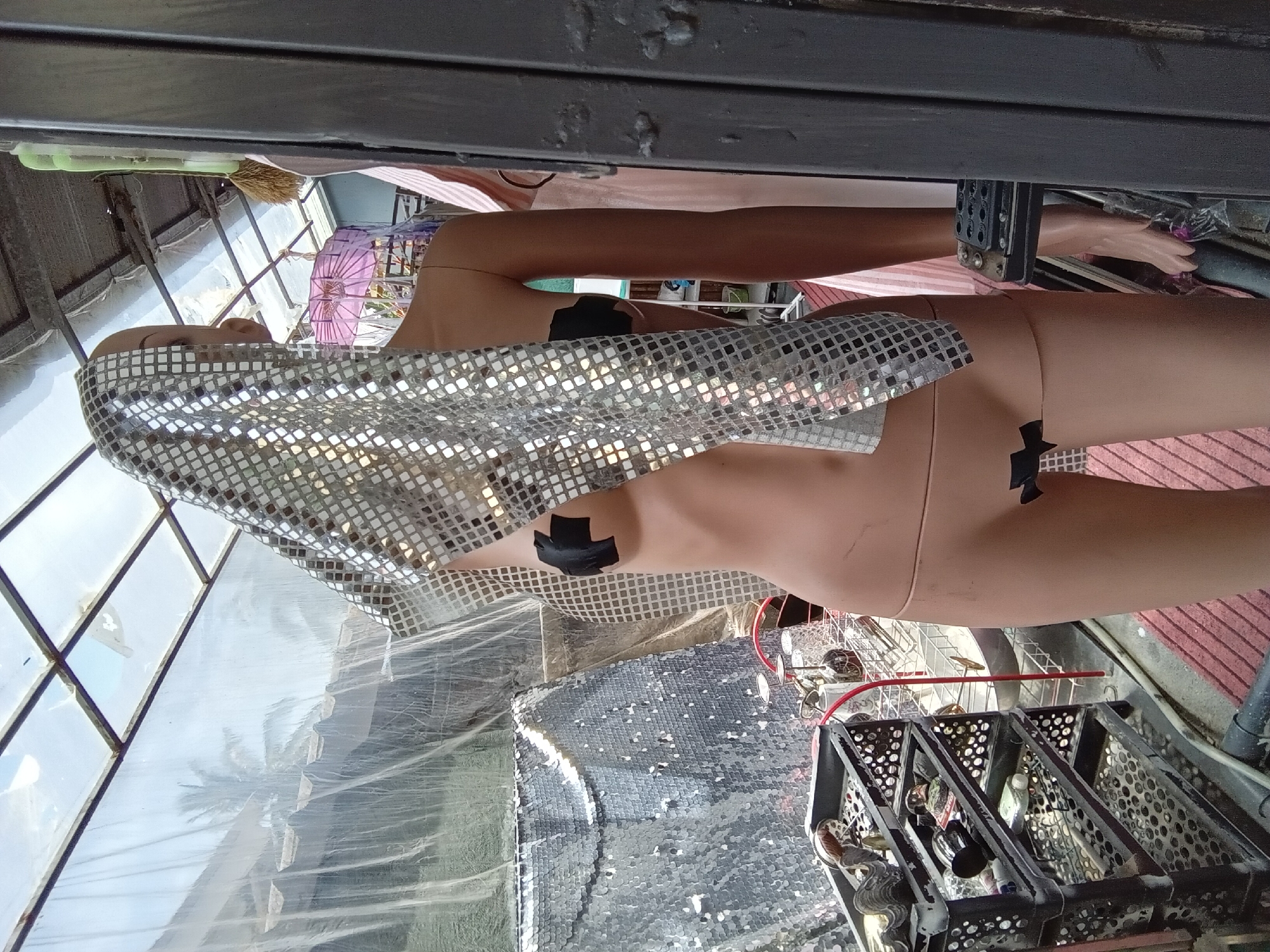 The image shows a mannequin wearing a sparkly silver top. The mannequin is positioned in what appears to be an outdoor or semi-outdoor setting, possibly a workshop or storage area, with a corrugated metal roof and a large window or glass wall. The background suggests a tropical location with palm trees visible through the glass. There is a red and white striped fabric draped behind the mannequin, and a pink umbrella can be seen in the distance. A black plastic crate filled with various items, including bottles and what looks like hair extensions, is in the foreground. The overall scene is cluttered, with a mix of materials and objects. The lighting suggests it is daytime, but the specific time of day and weather conditions are not clearly discernible. There is no visible text in the image.