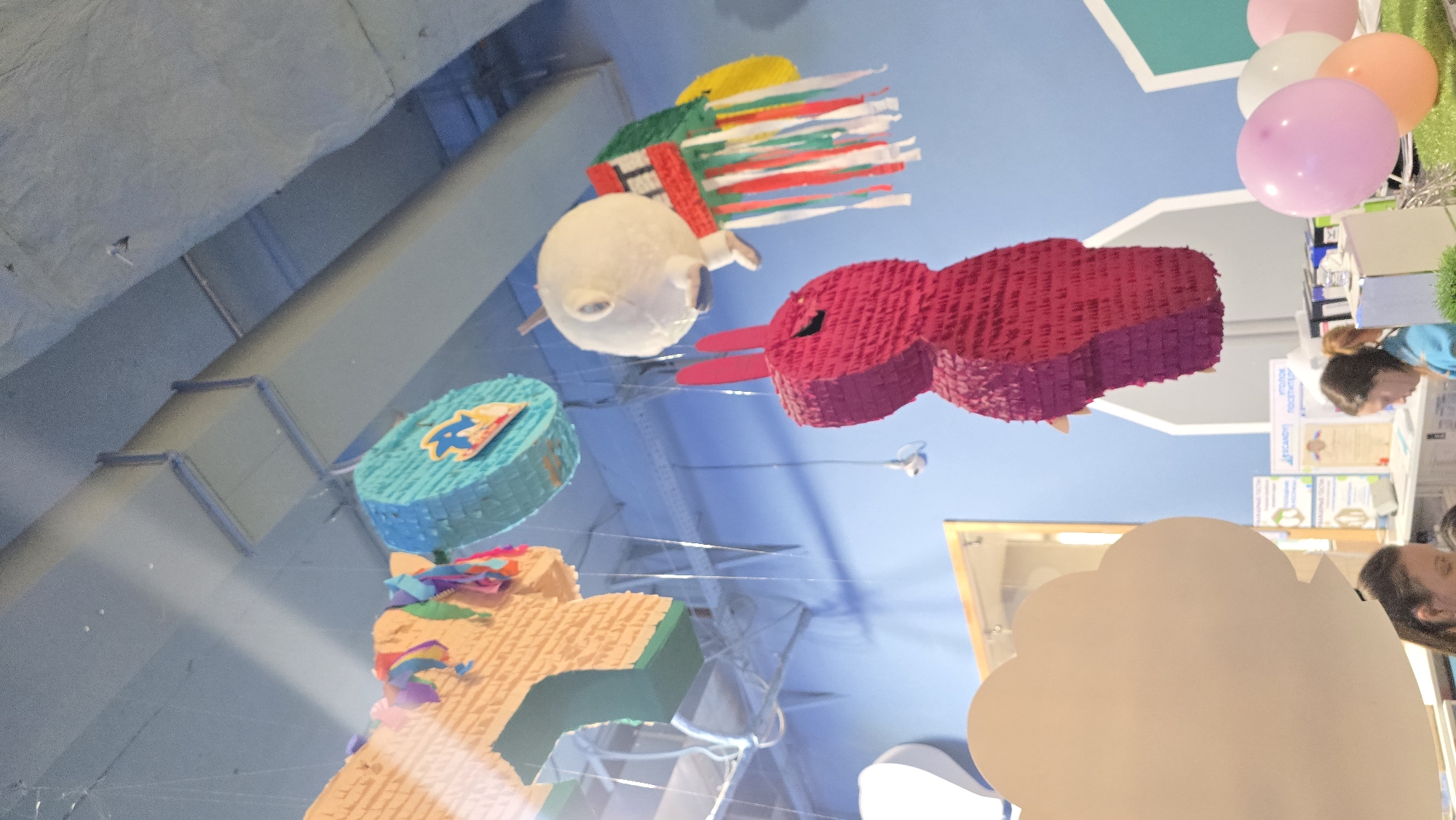 An indoor scene, likely a play center or event venue, features a bright blue wall and an industrial-style ceiling with exposed light blue-grey beams and ducts. Four distinct piñatas are suspended from the ceiling by thin transparent lines. On the far left, a light brown, multi-layered piñata, possibly shaped like an animal, is partially visible, adorned with colorful streamers. Next to it hangs a circular, teal piñata with a yellow and red emblem. To the right, a white, spherical piñata with a small dark face is attached to a green, red, and white fringed body, resembling a caterpillar, with a yellow disk at its rear. Prominently in the mid-foreground on the right is a large, dark pink, fringe-textured piñata shaped like a rabbit with tall ears.
In the lower right, a cluster of pink, white, and orange balloons rests on a textured green surface, possibly artificial grass. A large, white, cloud-shaped cutout partially obscures the bottom left corner of the image. Below this cutout, the forehead and hair of a woman are visible. Further to the right in the mid-background, two women are present at a counter or desk. One woman, with dark hair pulled back, wears a blue top and looks down, seemingly engaged with items on the desk. Behind her, another person's head is partially visible. On the counter, various objects, including bottles and what appear to be framed certificates or signs with text in Cyrillic script (e.g., "УГОЛОК ПОСЕТИТЕЛЕЙ" - Visitors' Corner), are arranged. A small white security camera is mounted on the upper right wall/ceiling structure. The scene is well-lit, with a diagonal beam of light visible on the ceiling in the upper left. The location is in Unknown, Unknown.