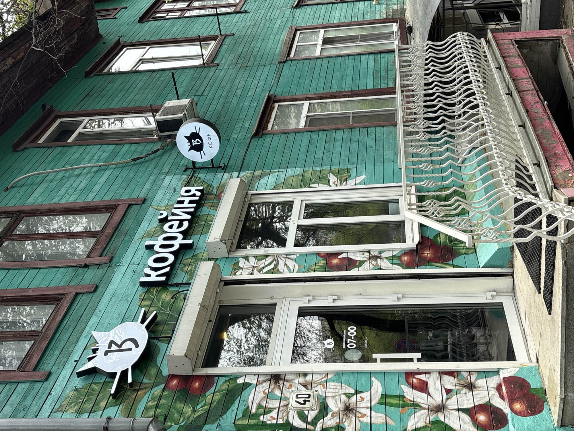 The image shows the exterior of a coffee shop called "Кофейня" (Coffee Shop) located in Khabarovsk, Russia. The building is painted turquoise with brown trim around the windows. A mural of coffee beans and white coffee flowers adorns the lower portion of the building's facade. A sign with a black cat silhouette and the letter "B" is displayed above the entrance. The shop's business hours are marked as "07-00". The building's address number "40" is visible near the bottom. Several windows are visible, reflecting the trees outside. An air conditioning unit is mounted above one of the windows. A white decorative wrought iron railing is attached to the building on the right side of the frame.