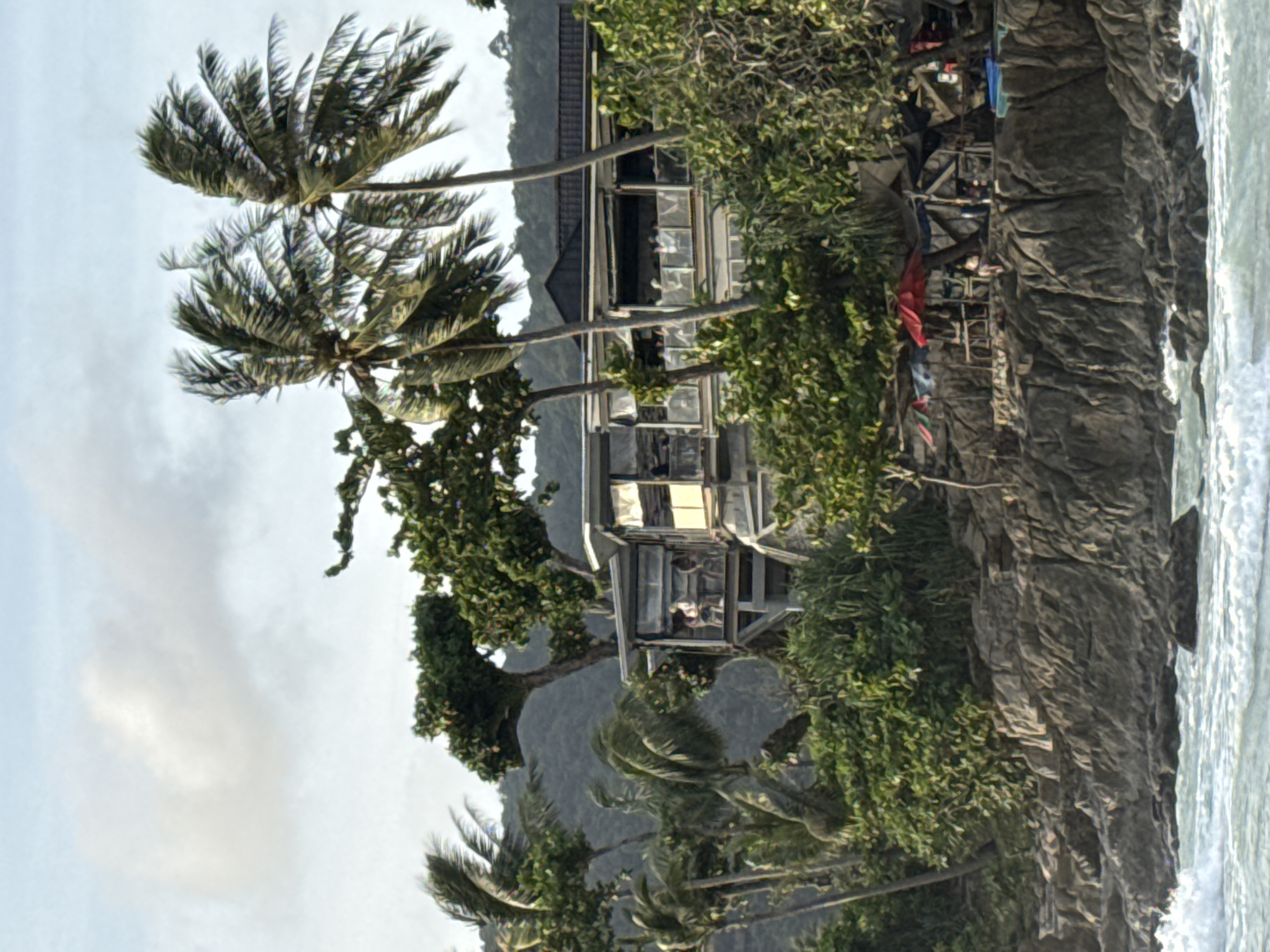 In Patong, Thailand, a restaurant is situated on a rocky shoreline, surrounded by lush vegetation and swaying palm trees. The building has multiple levels with large windows, revealing patrons seated at tables inside. In the foreground, waves crash against the rocks. The sky above is overcast with scattered clouds. To the right of the restaurant, a small cluster of beach umbrellas and makeshift shelters suggests a relaxation area.
