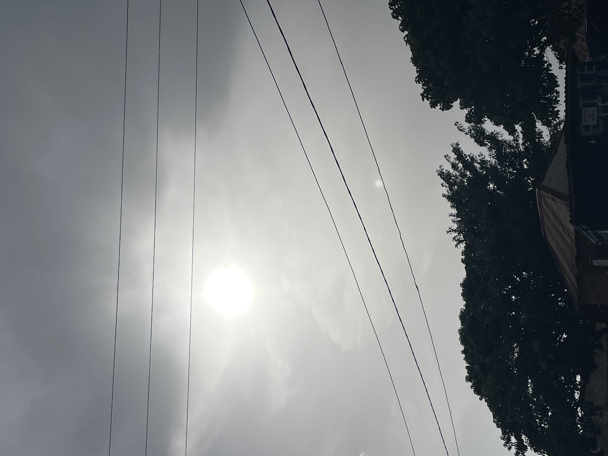 The image shows an upward-angled view of a cloudy sky with visible power lines. The sun is obscured by the clouds but still brightly visible. To the right, there are dark trees and a building, likely residential. The location is presumed to be Durimi, Nigeria, though this is not discernible from the image itself. There are no visible people or interactions.